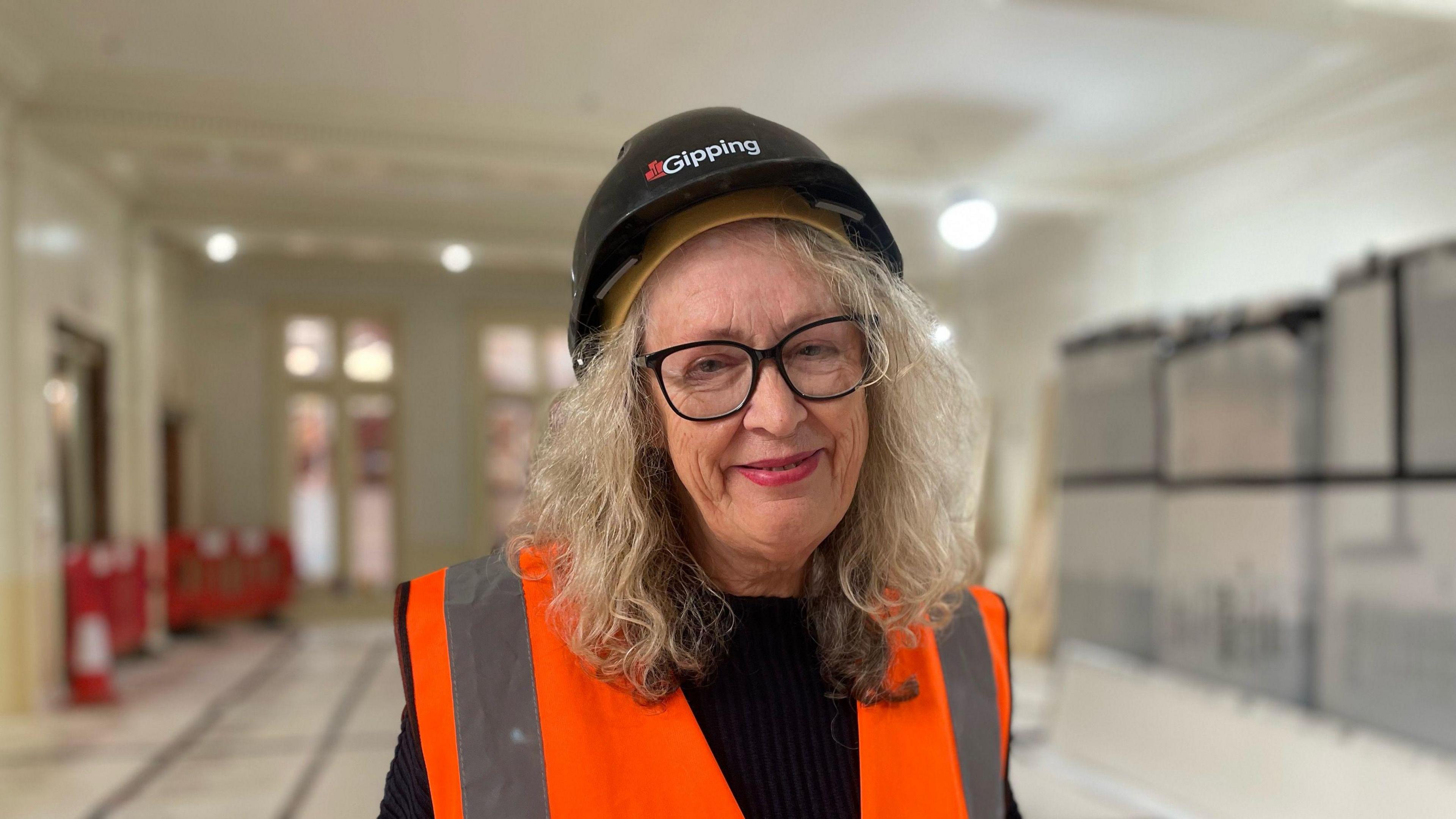 A photo of Carole Jones. She is facing the camera and smiling. She is wearing a black safety helmet and an orange high-vis jacket. She has a black top on underneath and she is wearing black glasses. She has shoulder-length blonde hair. She is standing in one of the museum's rooms which is blurred behind her.