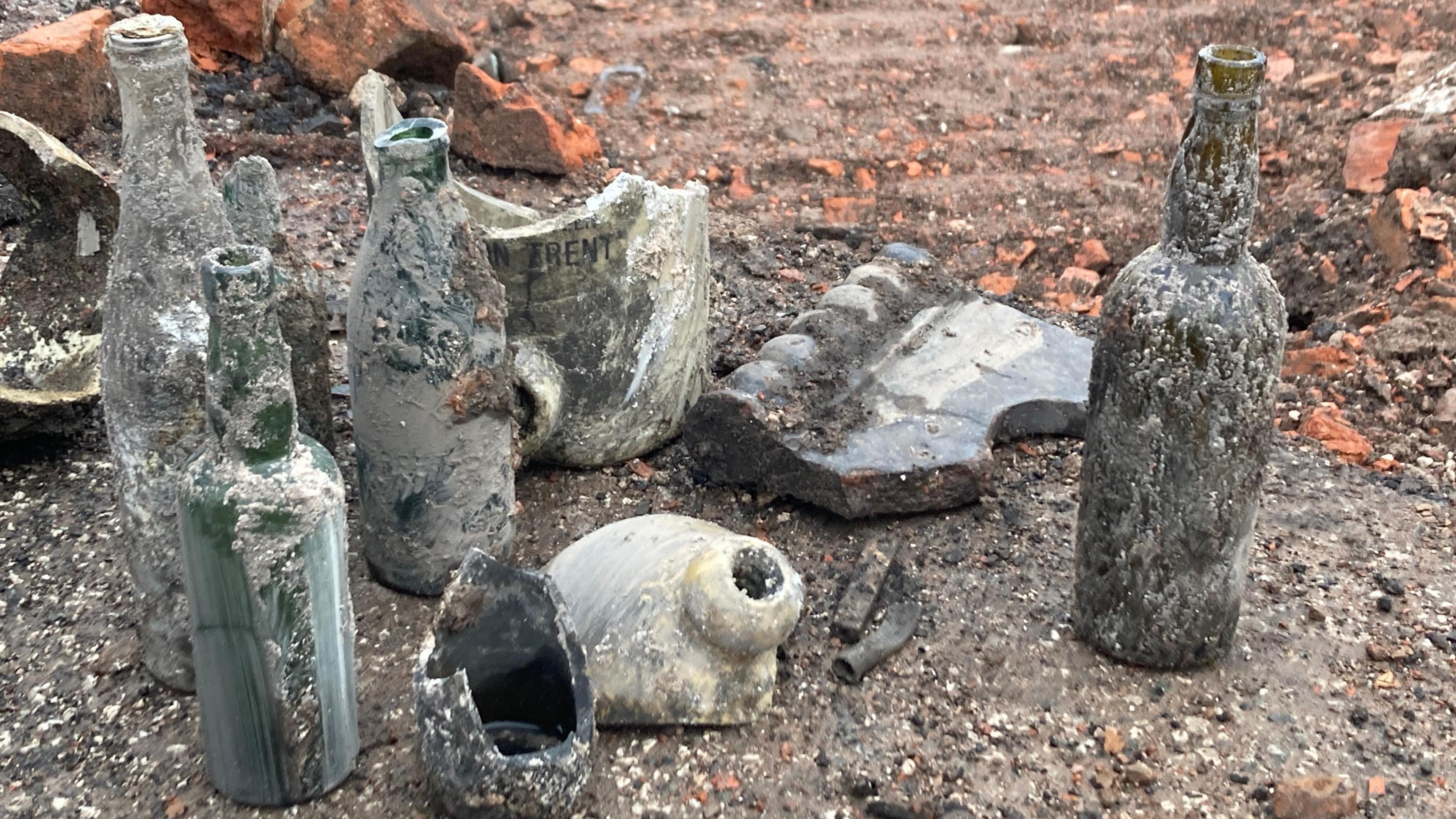 Four old bottles, encrusted in dirt are lined up on the floor next to pieces of broken pottery