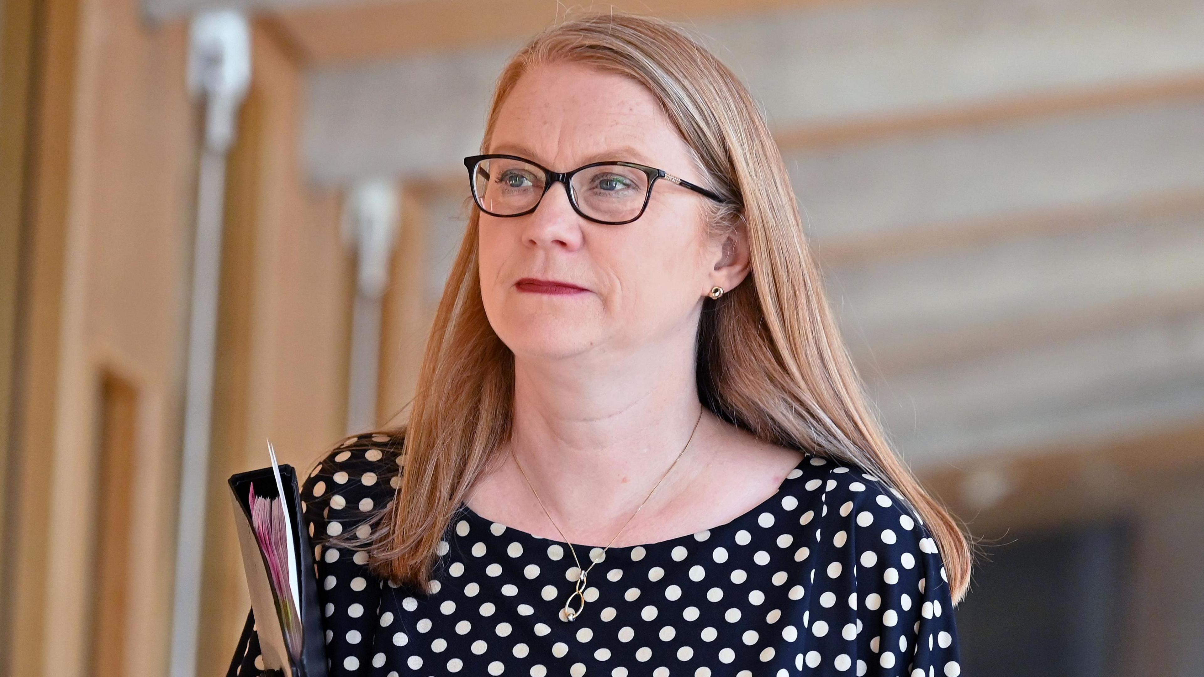 Shirley-Anne Somerville - a a woman with glasses, shoulder length auburn and wearing a black dress with white polka dots - walks in a corridor at Holyrood while carrying a folder