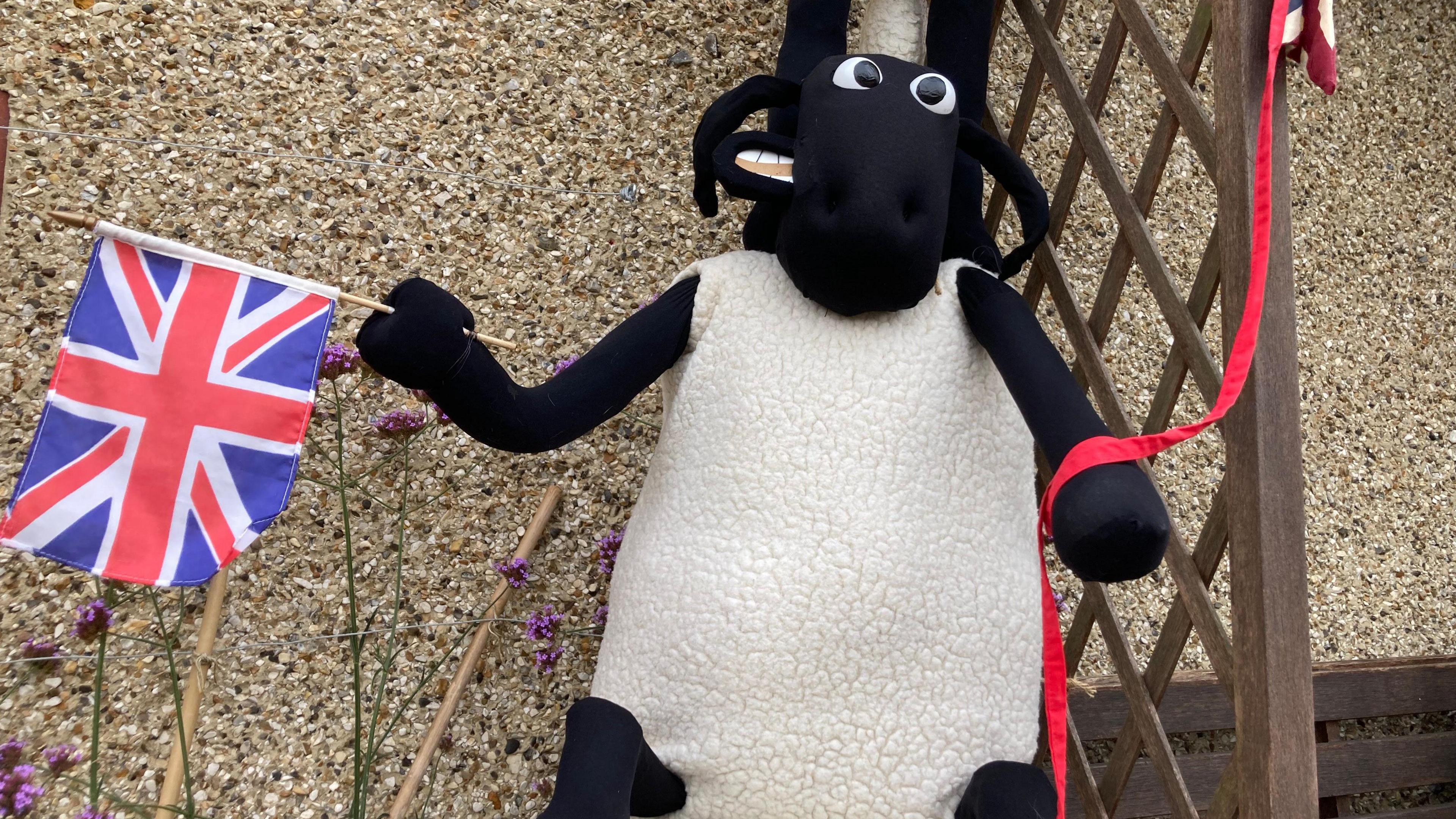 A scarecrow of Shaun the Sheep. The sheep has a black face and legs and a white fluffy body. The scarecrow is holding a Union Jack.