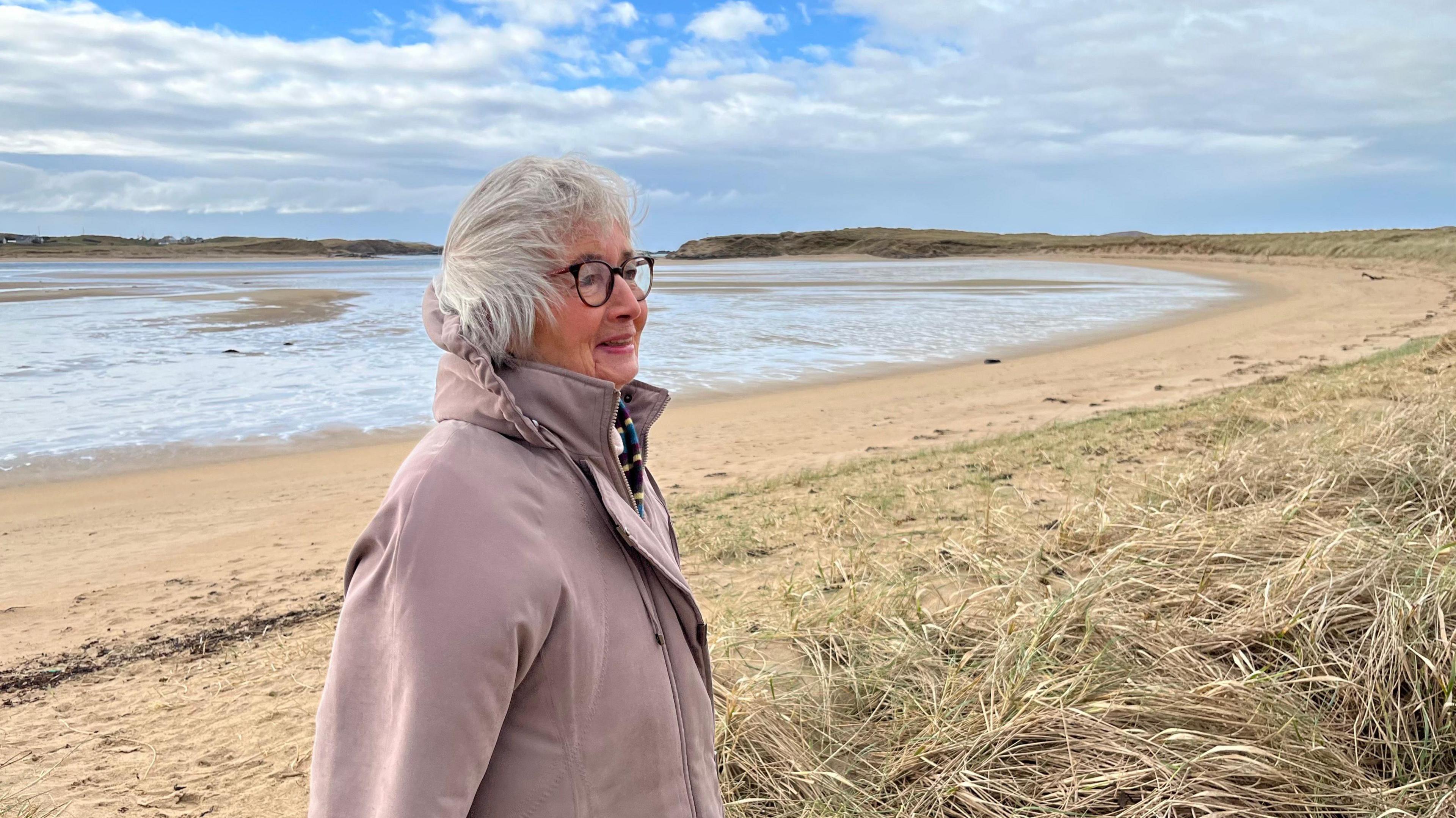 Bríd Rodgers on a windswept sandy beach in County Donegal  She is smiling and looking into the distance.  She has grey hair and glasses and is wearing a stone-coloured overcoat with a hood. 