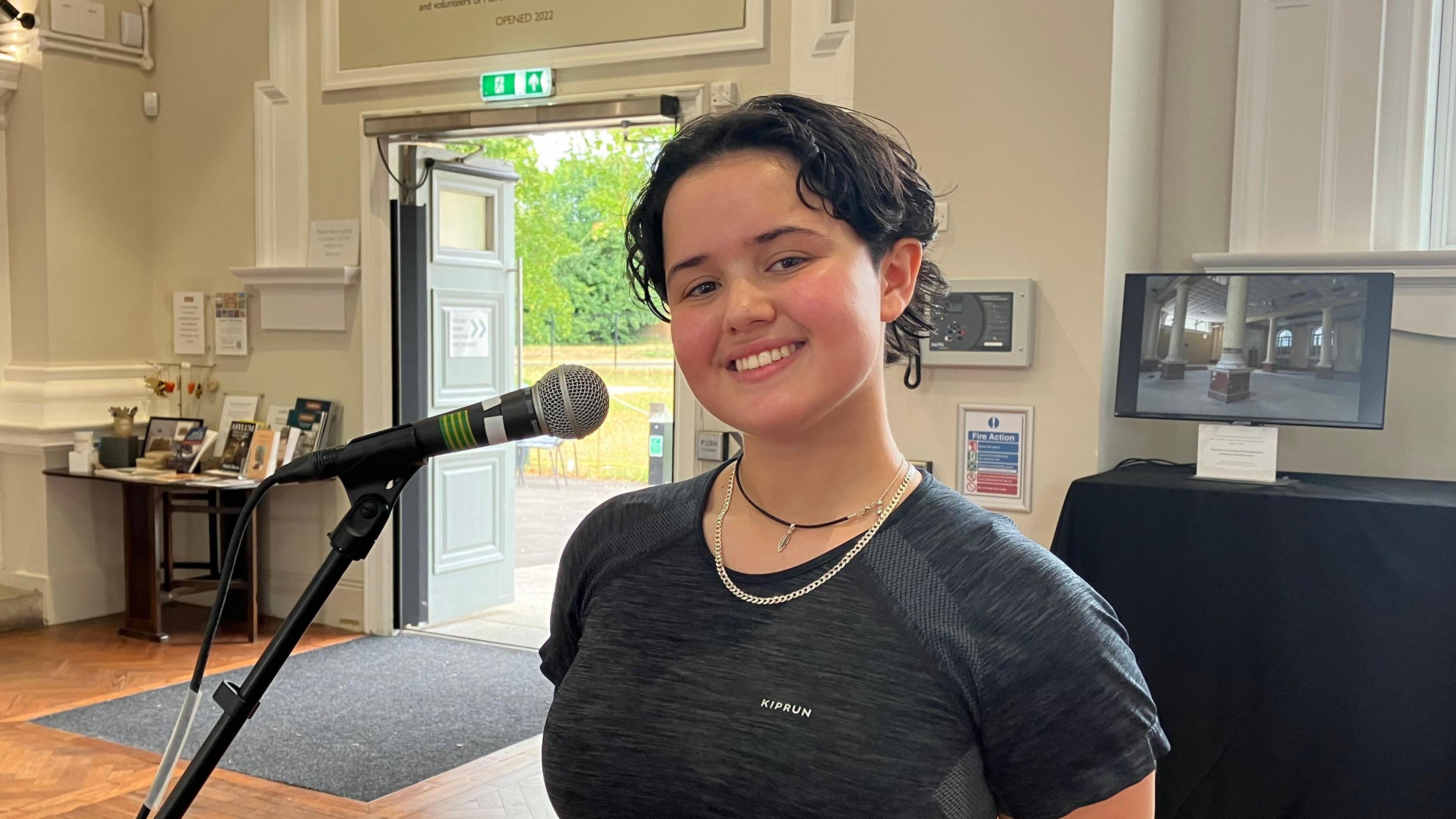 Sara May looks at the camera and smiles. She is standing at a microphone and wearing a black t-shirt.
