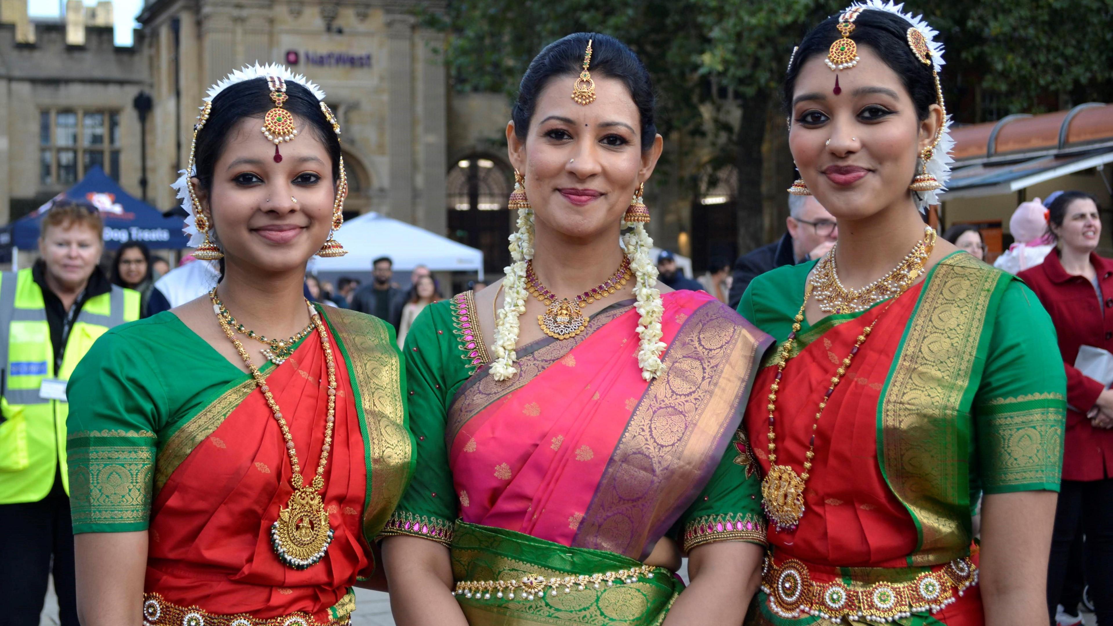 Three women dressed in colourful sarees and gold jewellery with flowers in their hair, smile and look directly at the camera. 