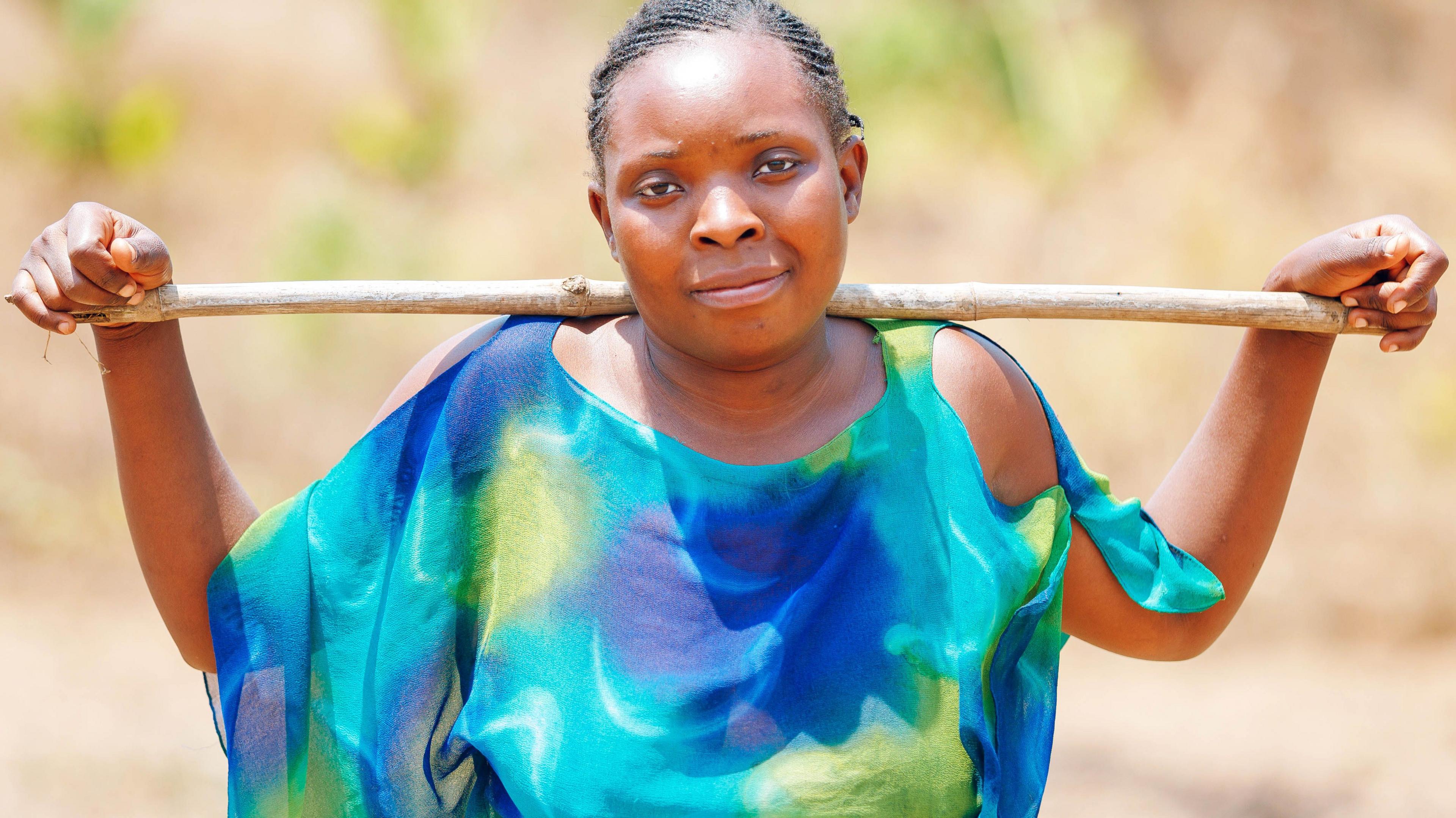 A portrait of Abigail Namtowe, 28, a farmer in Twalima village. She smiles modestly at the camera and rests a long wooden stick along her shoulder blades, bringing her hands up to rest on them. She is wearing a bright blue and green flowy top. 