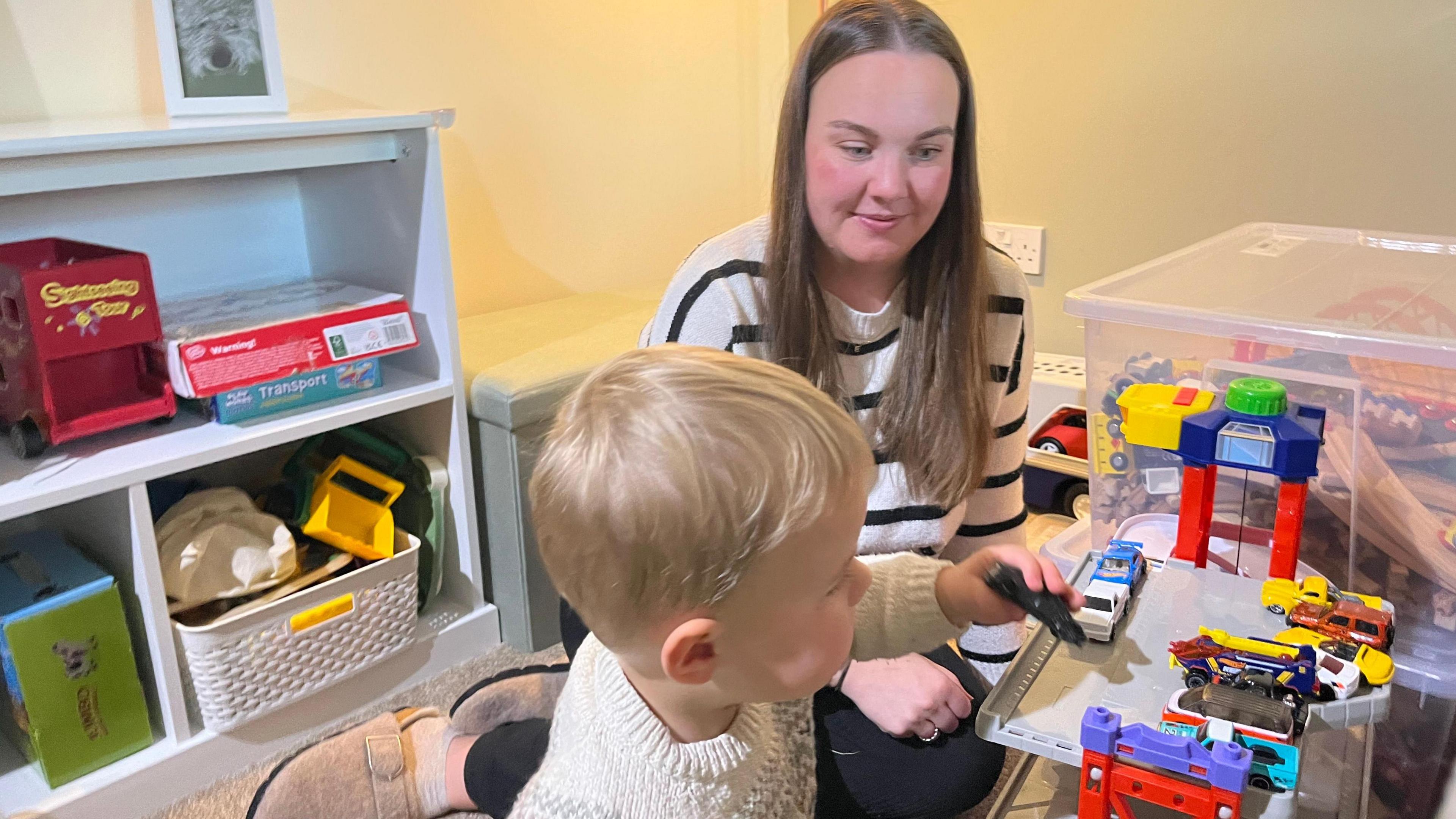 Fiona watches her son Euan play with his toy cars. She is sat on the floor of a cluttered bedroom which has toys strewn all over the floor. She has long brown hair and is wearing a white jumper with blue stripes.