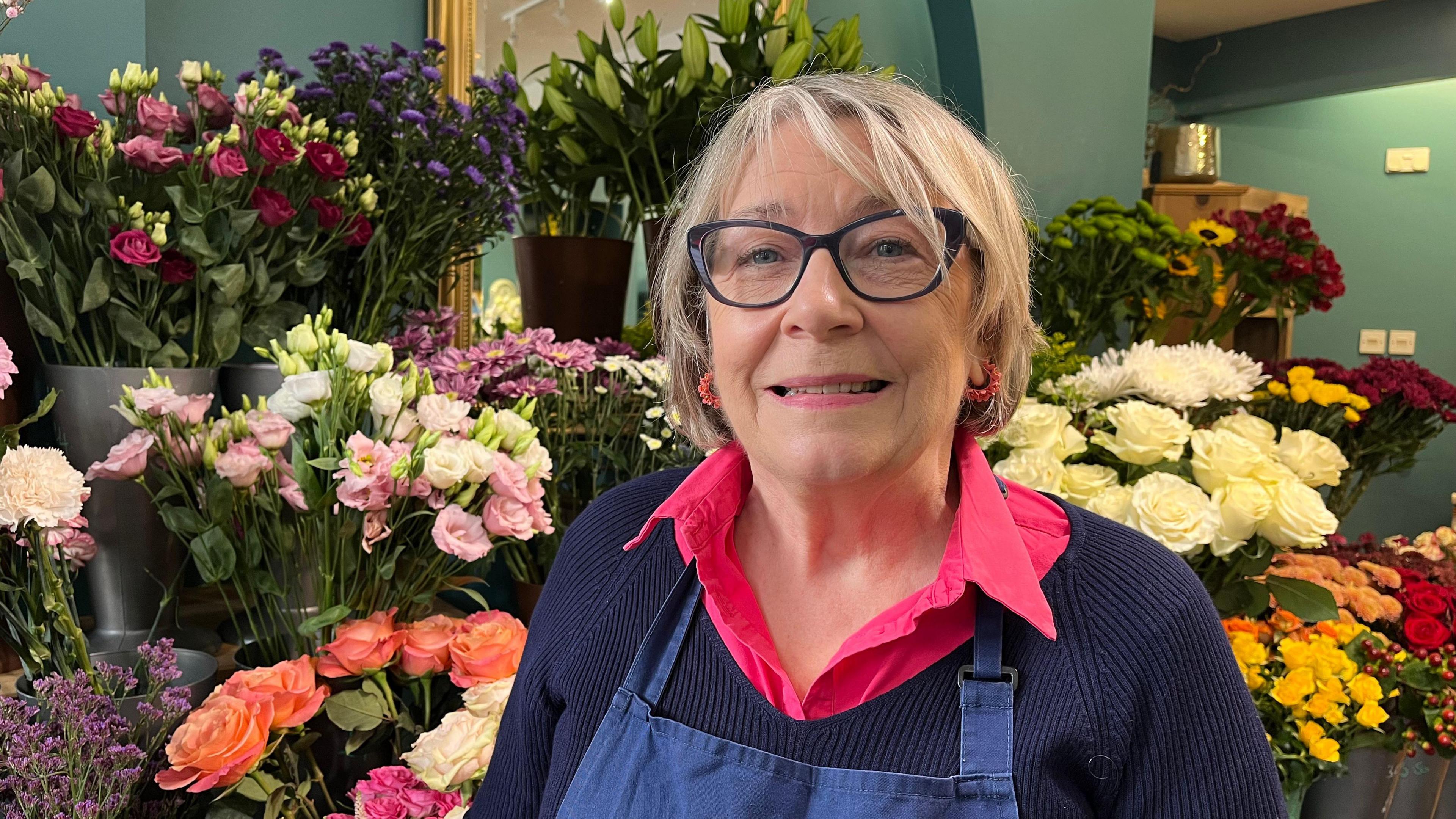 Catherine pictured looking into the camera and smiling. She's has blonde and grey hair and is wearing a bright pink collared shirt, a navy knitted jumper, and a navy blue apron. She's stood in front of three rows of buckets of flowers - mainly roses - in colours including white, apricot, dark red, and yellow. The walls of the room are teal blue.