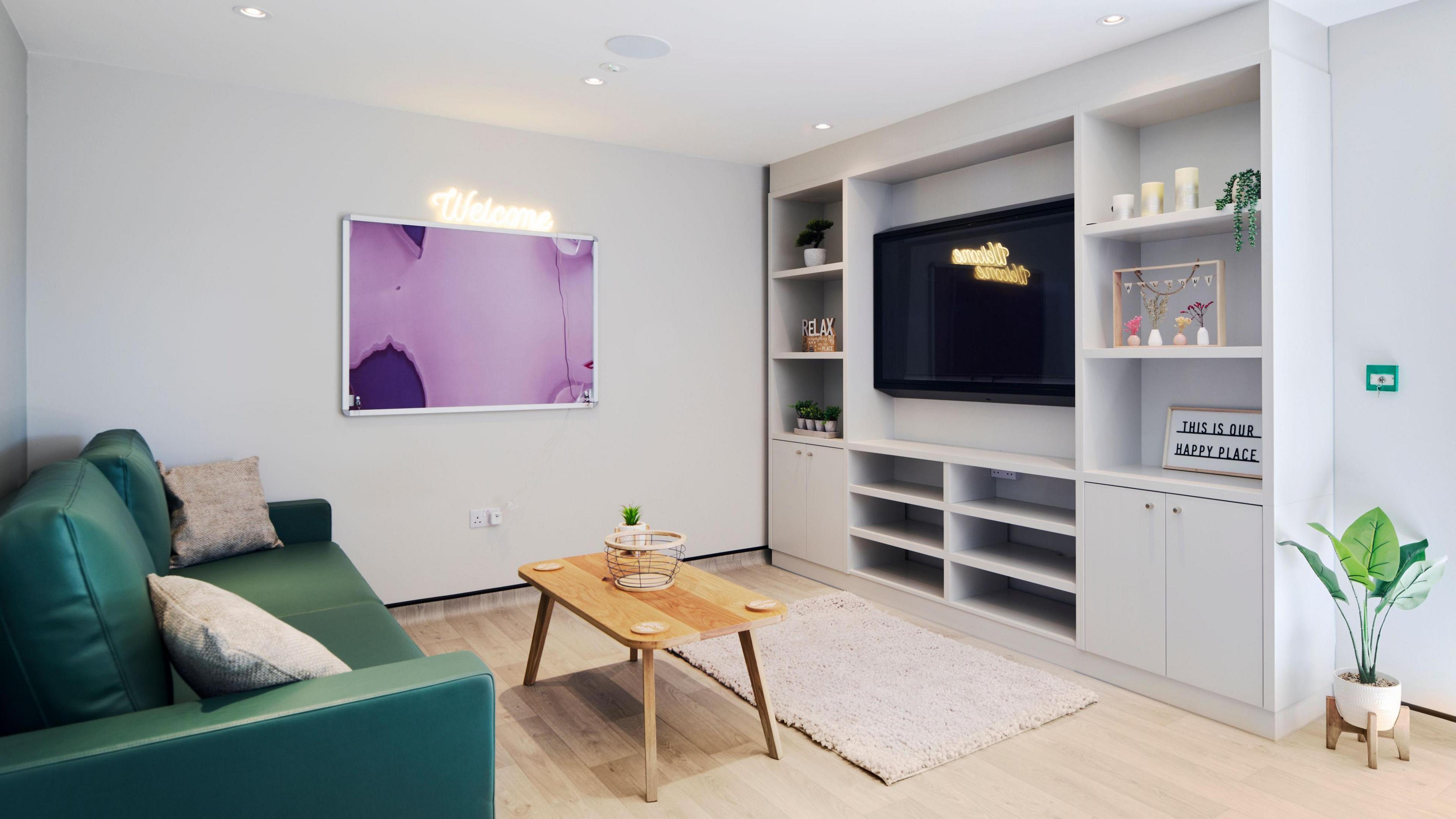 A green sofa with cushions sits opposite a TV unit with candles and pictures on the wall with a cream rug and coffee table.