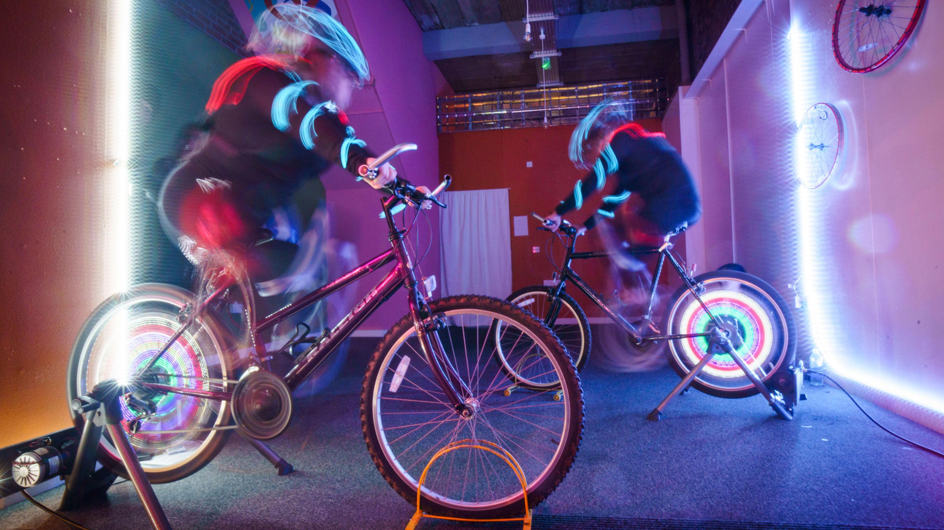 A blurred photo suggesting motion of two people on stationary bikes which have multicoloured lights in their spokes