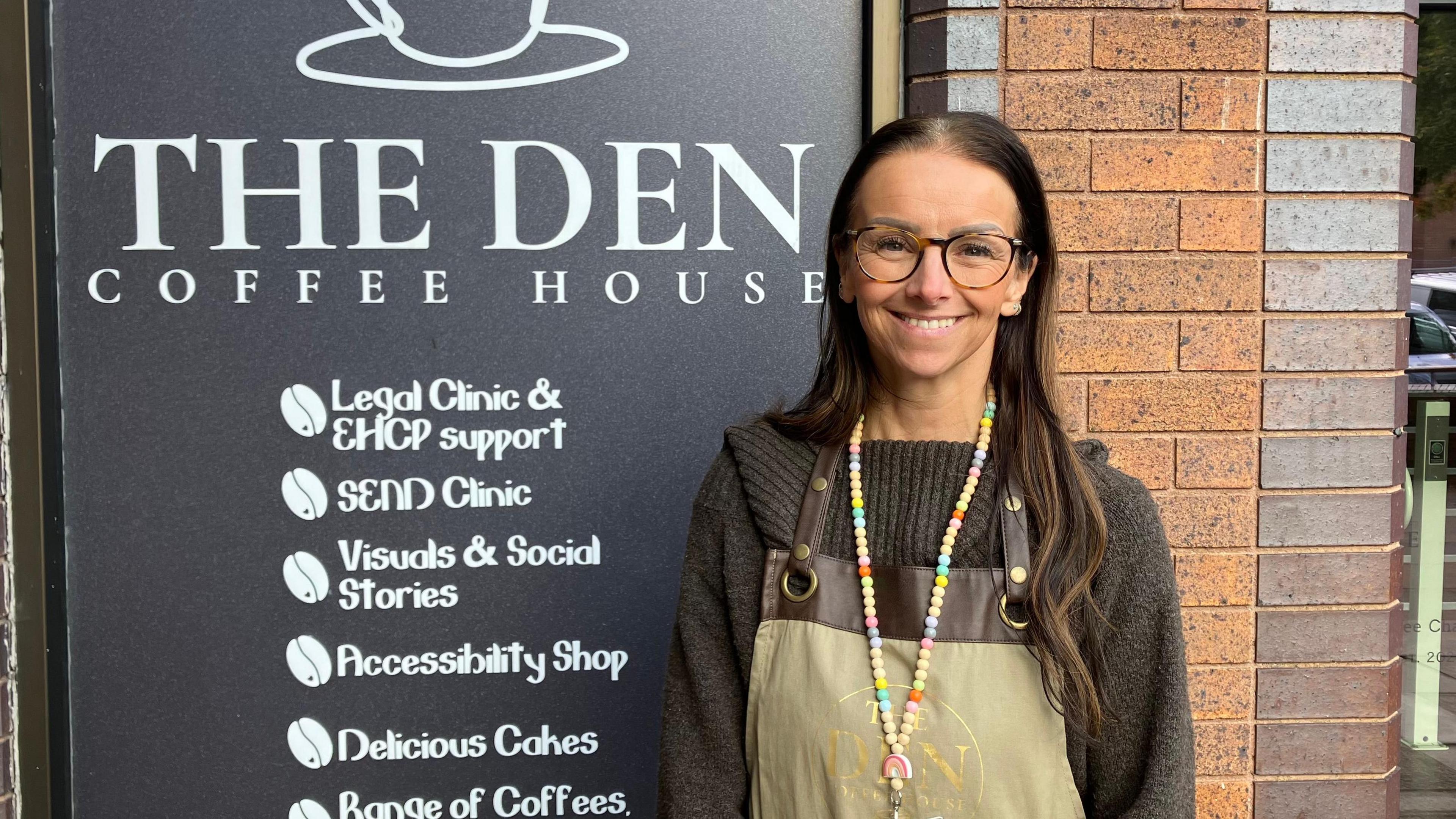 Claire Naylor is wearing a black jumper with an apron, and glasses, standing in front of a large sign for The Den Coffee House which lists its services. Services include a Send clinic, an accessibility shop and support with education, health and care plans.