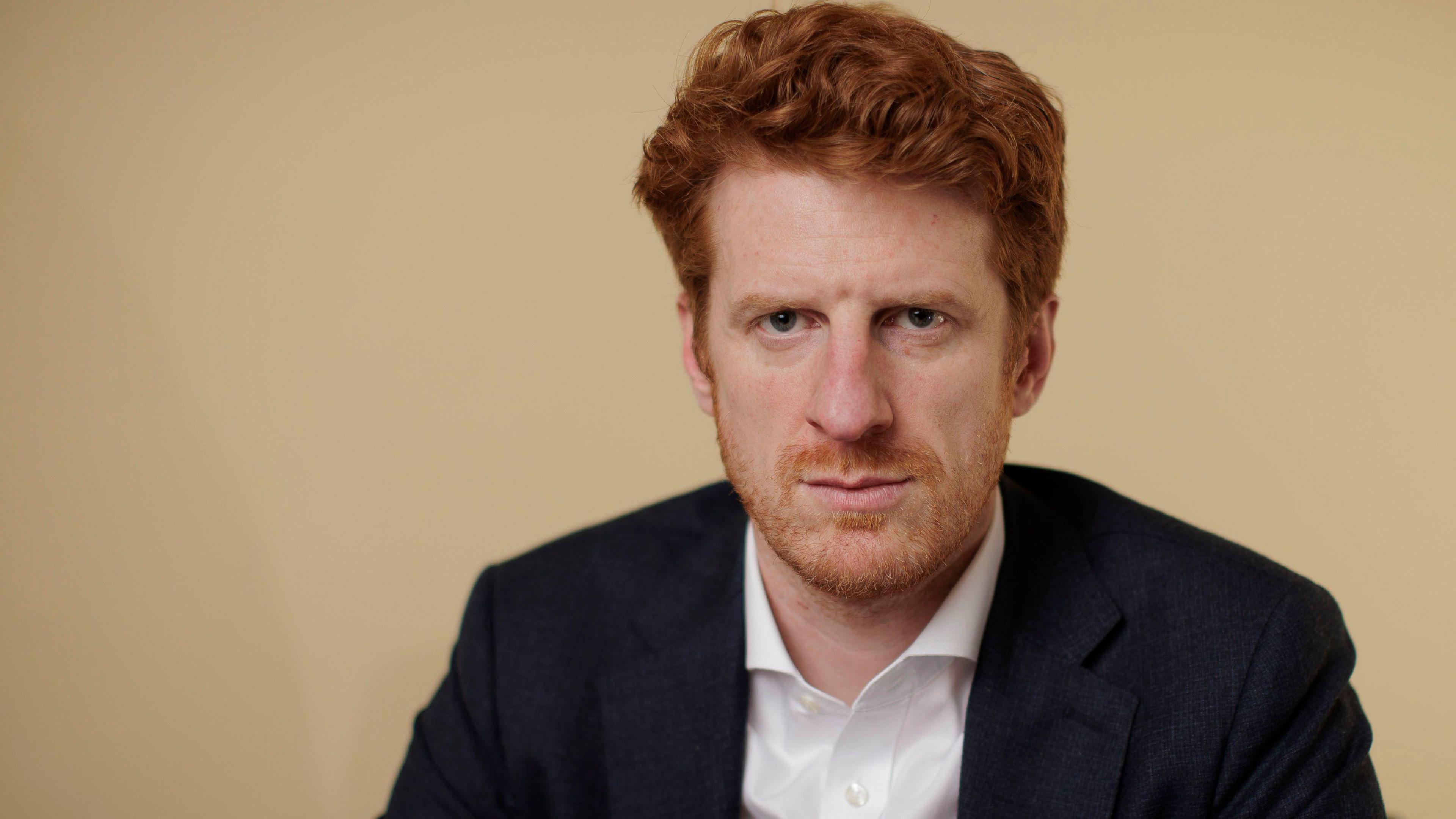 A man with red hair and beard, he is looking towards the camera. He is wearing a navy suit.