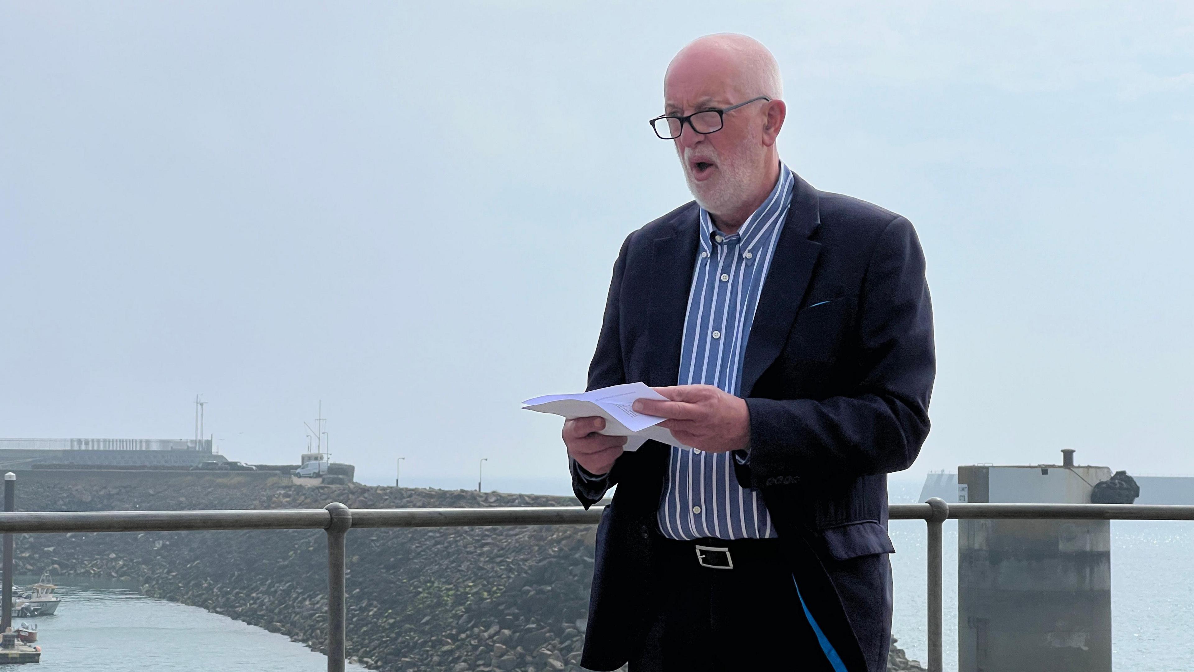 Deputy Luce delivers a speech as he holds some paper in front of him and behind is the St Helier Marina