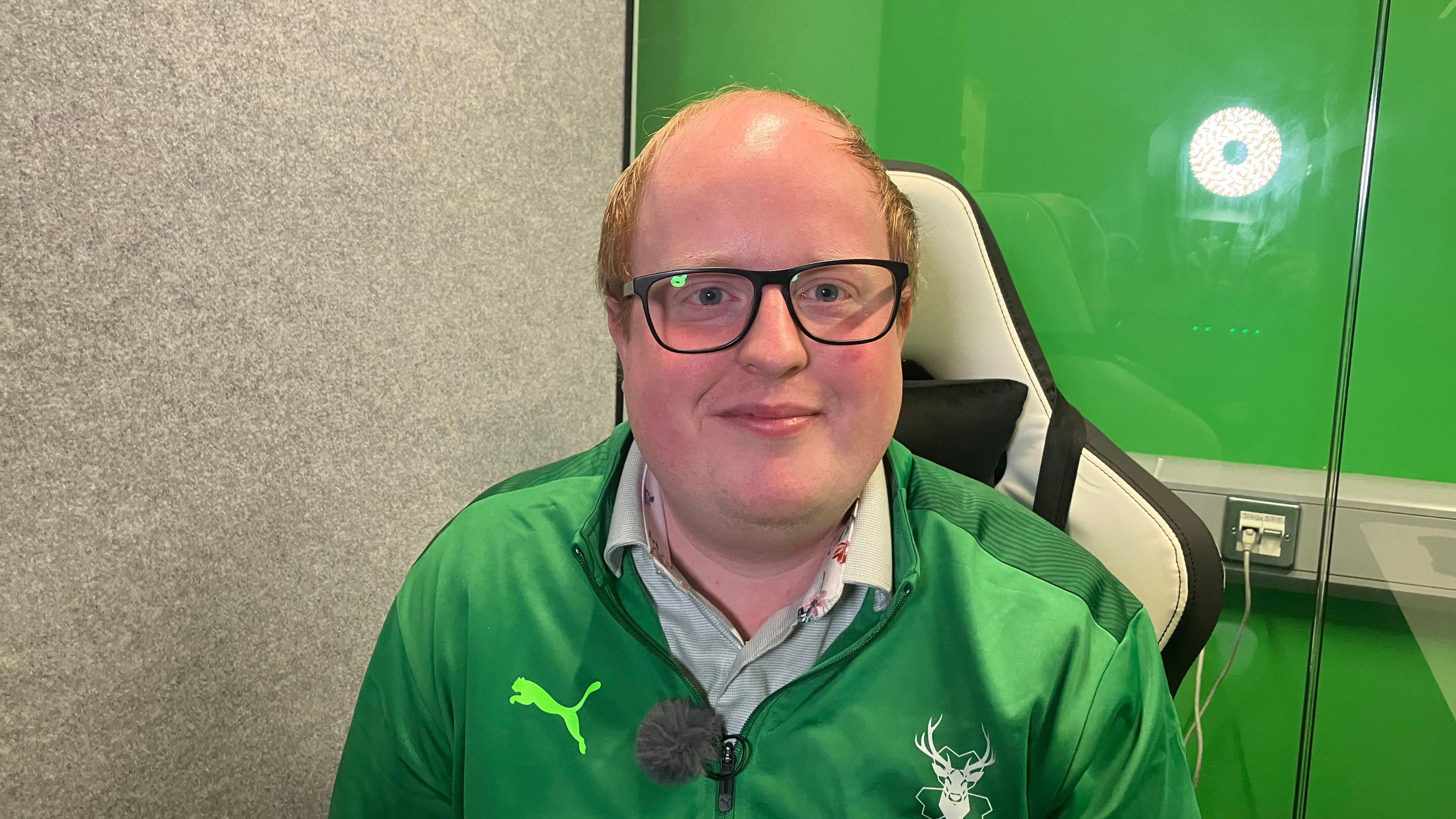 Michael Smyth smiles. He has glasses and ginger hair. He is wearing a green Puma zip up jersey. Behind him there is a green wall.