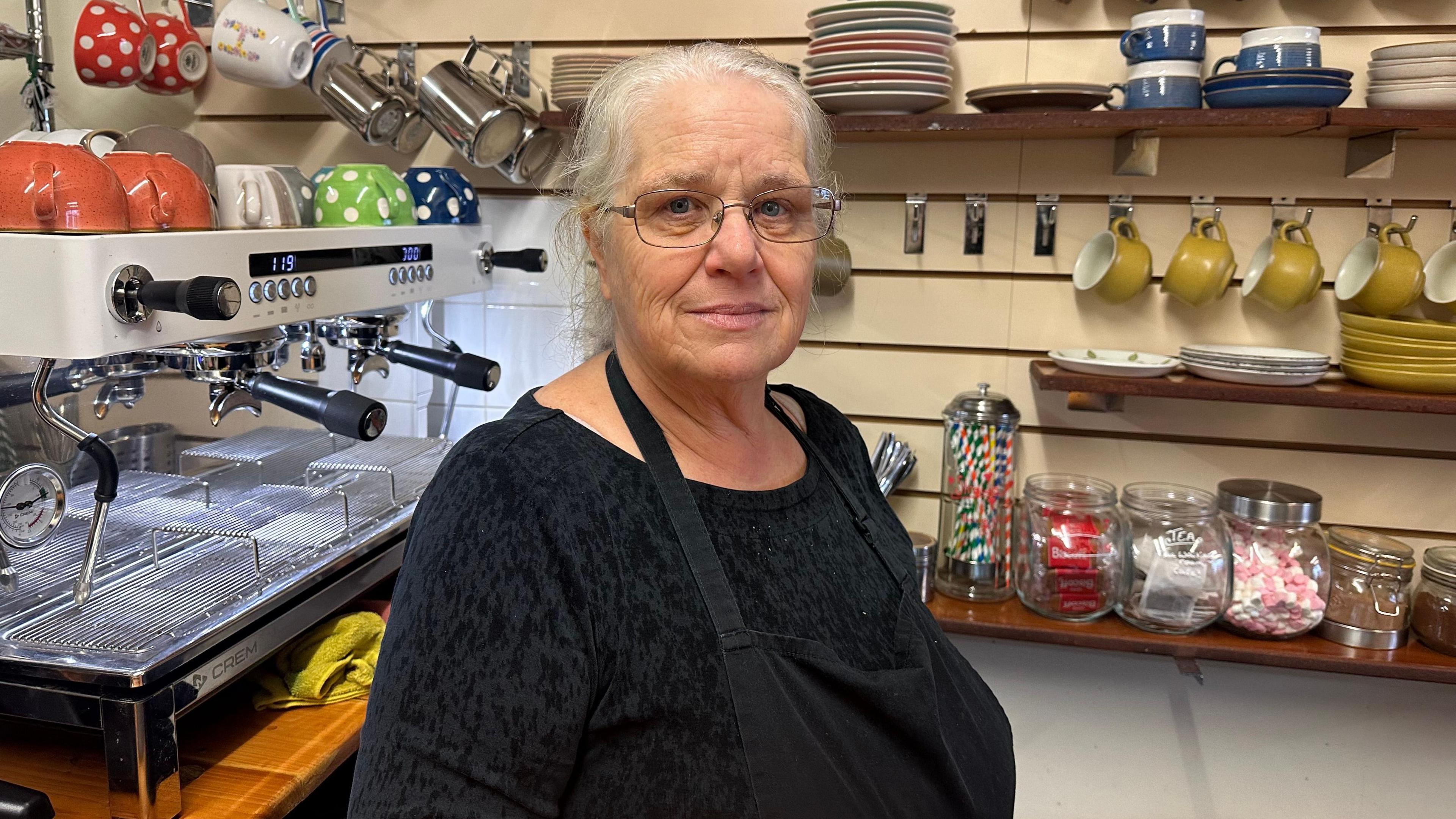 Carol Bowie wears glasses and has her light white hair in a ponytail. She is wearing a black top with a black apron and a barista coffee machine is in the background, along with shelves of hanging mugs. 