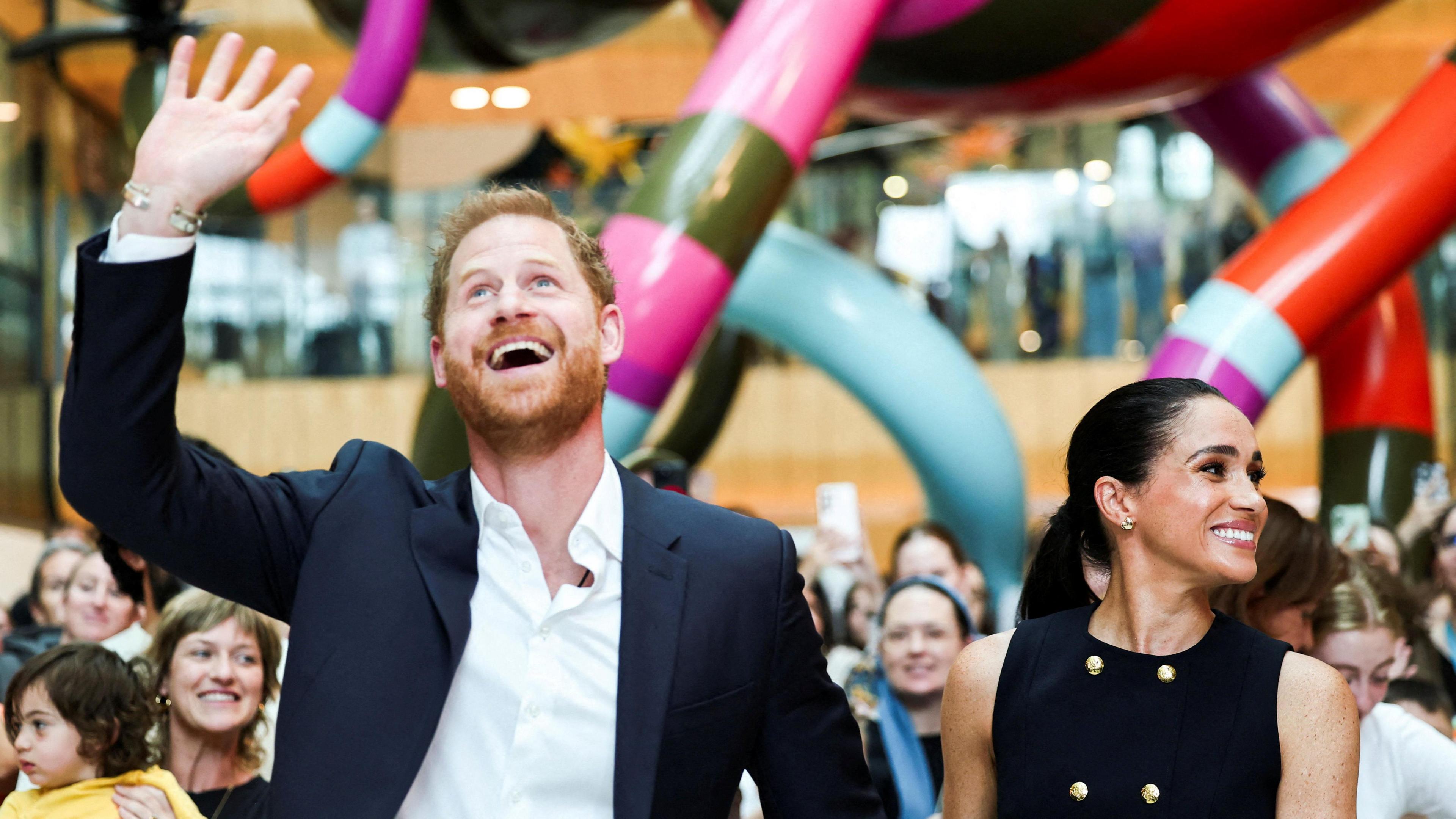 On the left, Prince Harry smiles, looks up and waves his right hand in the air, and on the right, Meghan smiles and looks to her left in front of a large crowd and a large colourful scuplture