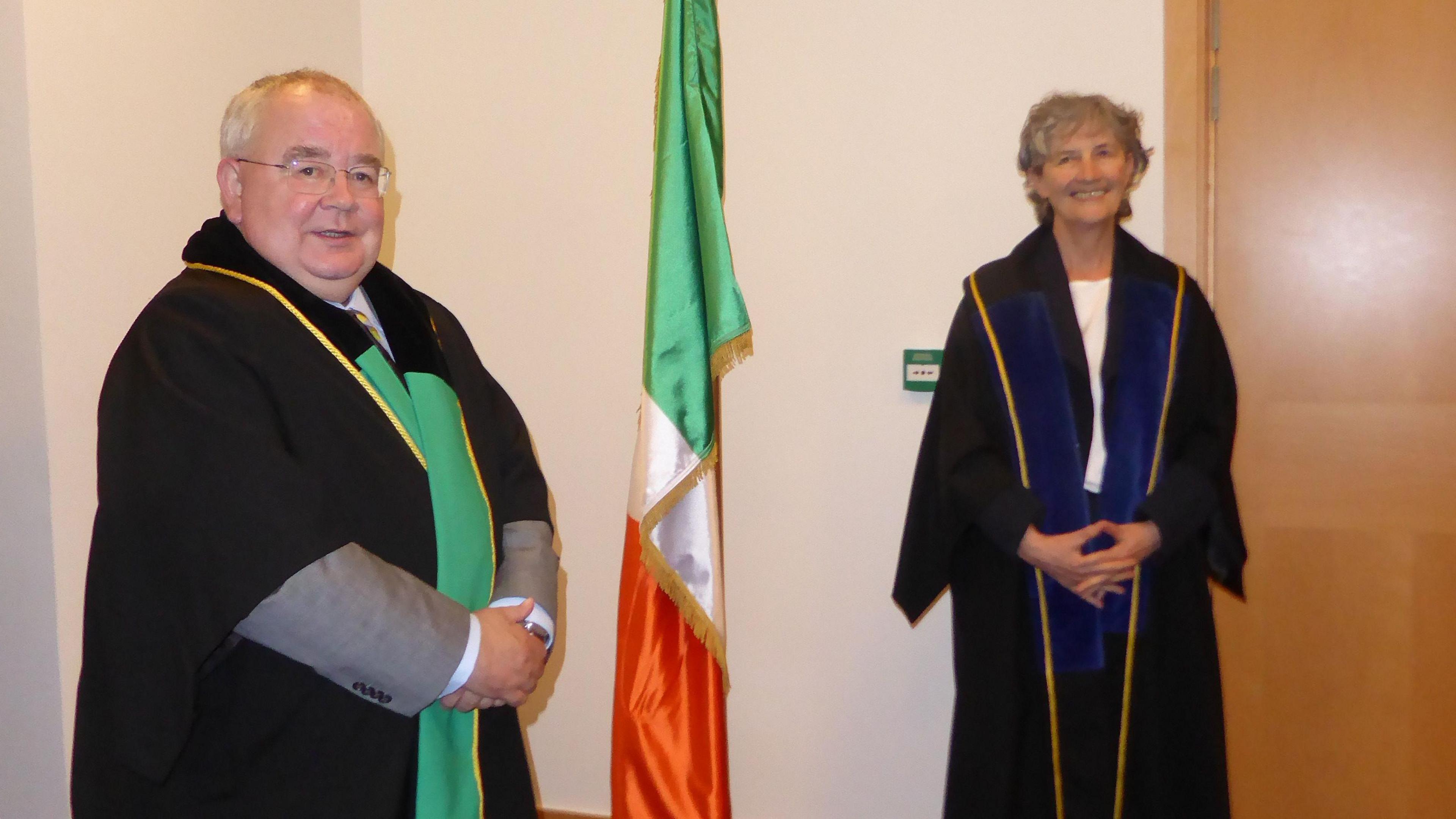 Catherine Connolly wearing a black robe with navy and gold detail following her election as Deputy Speaker of the Irish parliament.  She is pictured with Ceann Comhairle (Speaker) Seán Ó FearghaÃl who is wearing a black robe with green and gold detail.  The Irish flag stands on a tricolour between them