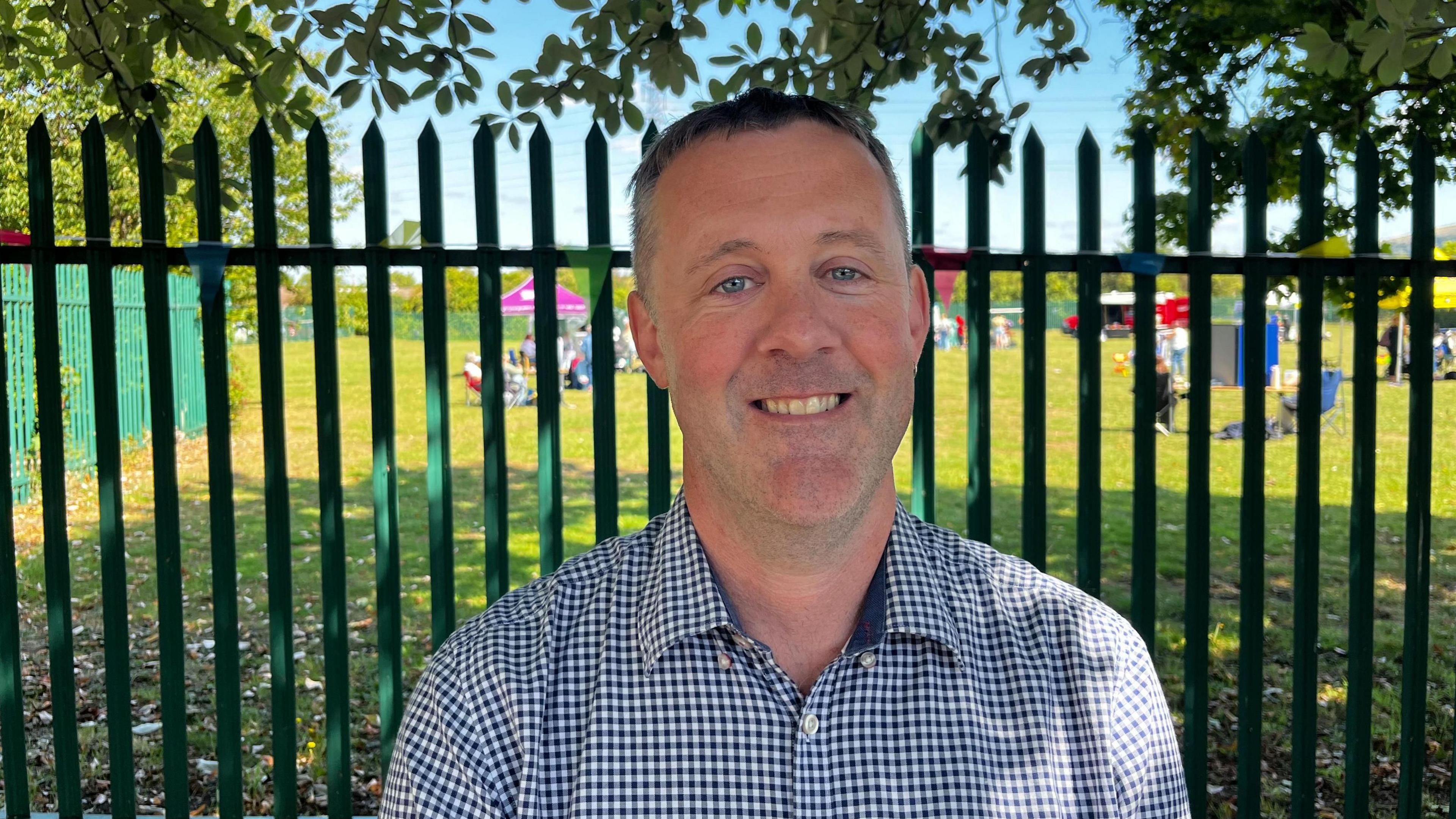 The image shows a man standing in front of a green fence wearing a blue and white checked shirt.