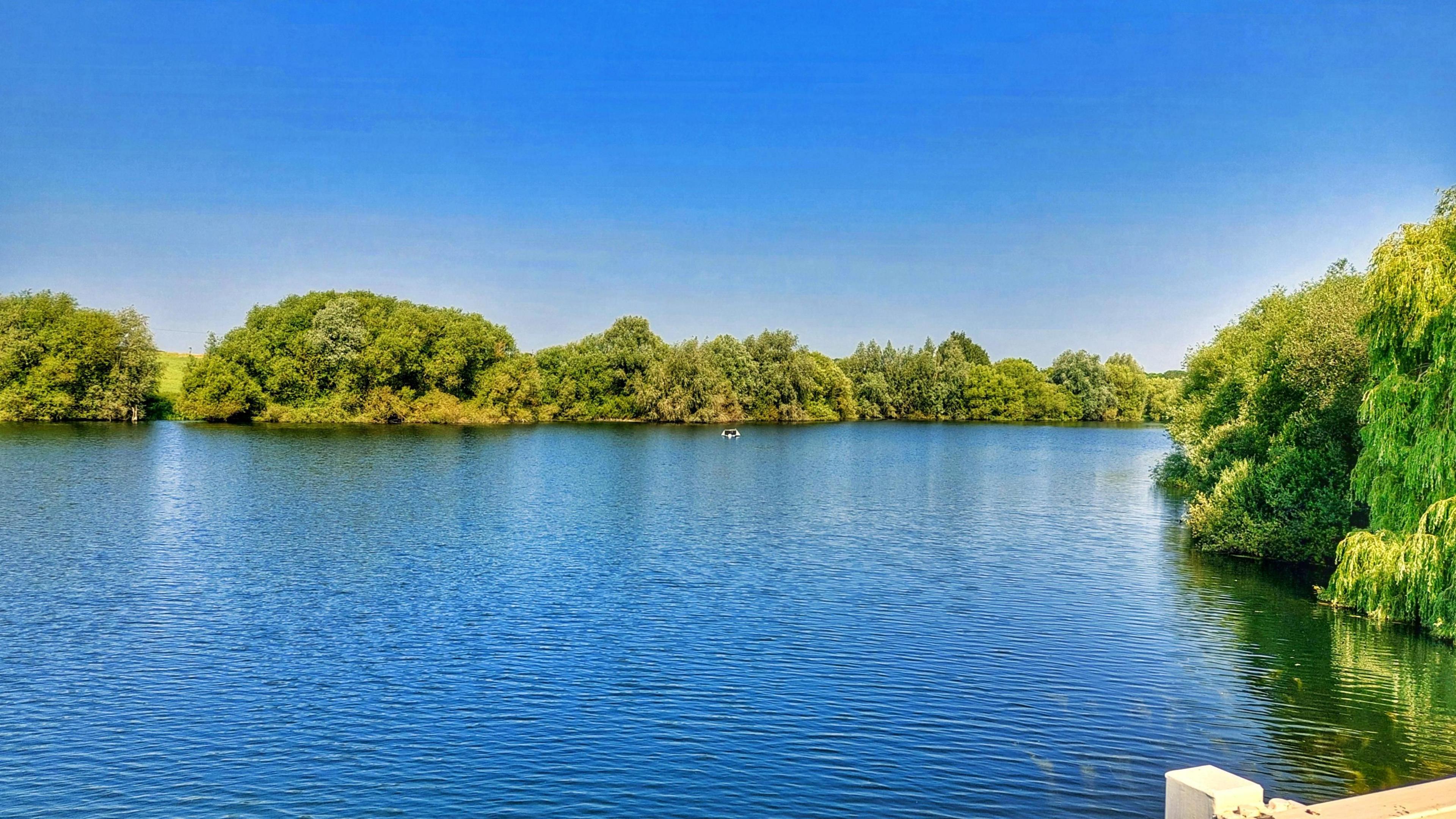 A view of Tattingstone reservoir on a summer's day. Green trees line the edge of the reservoir.