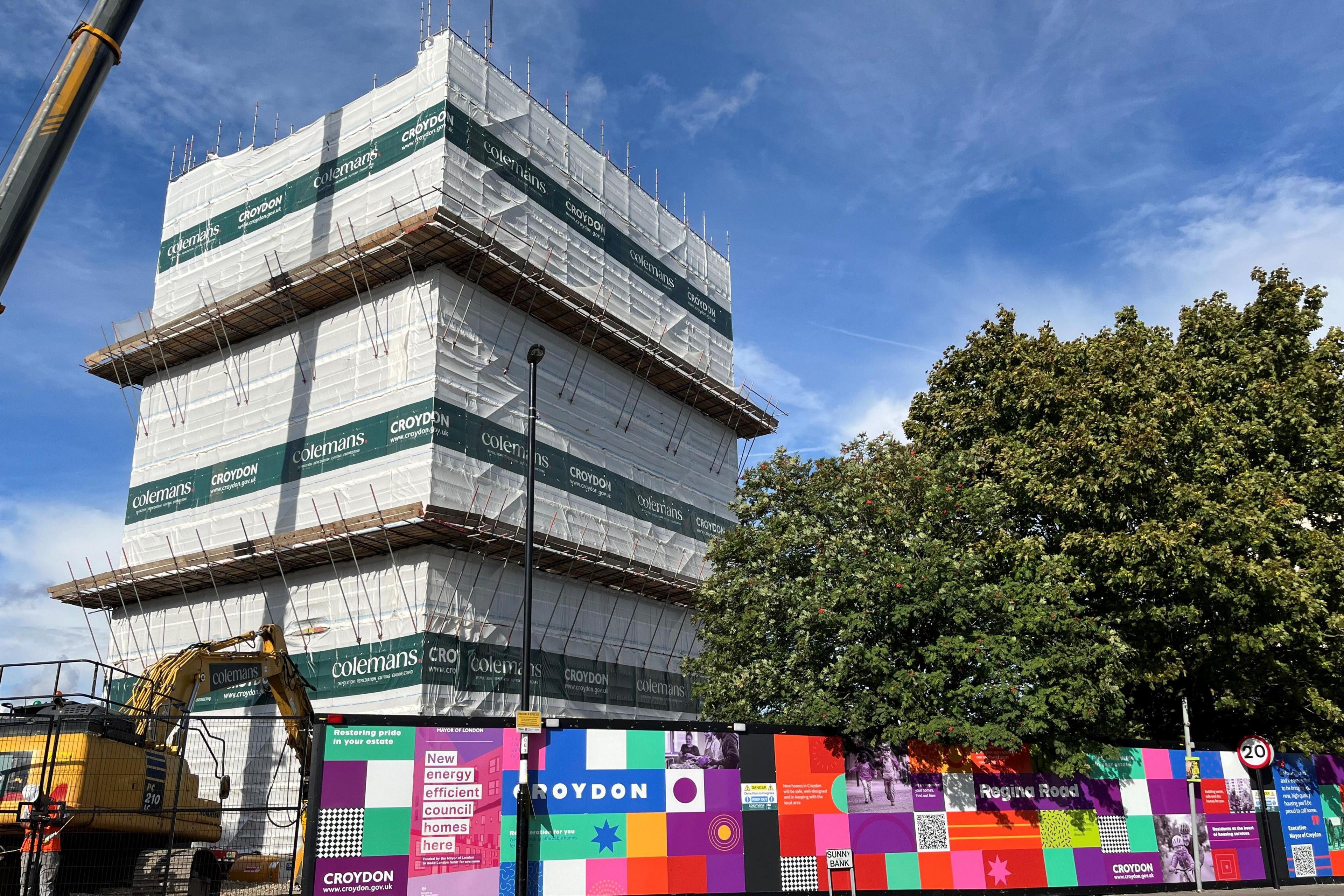 Block of flats under demolition covered in tarpaulin on the Regina Road estate in South Norwood, with yellow construction vehicle and colourful hoardings with Croydon Council signage in the foreground.