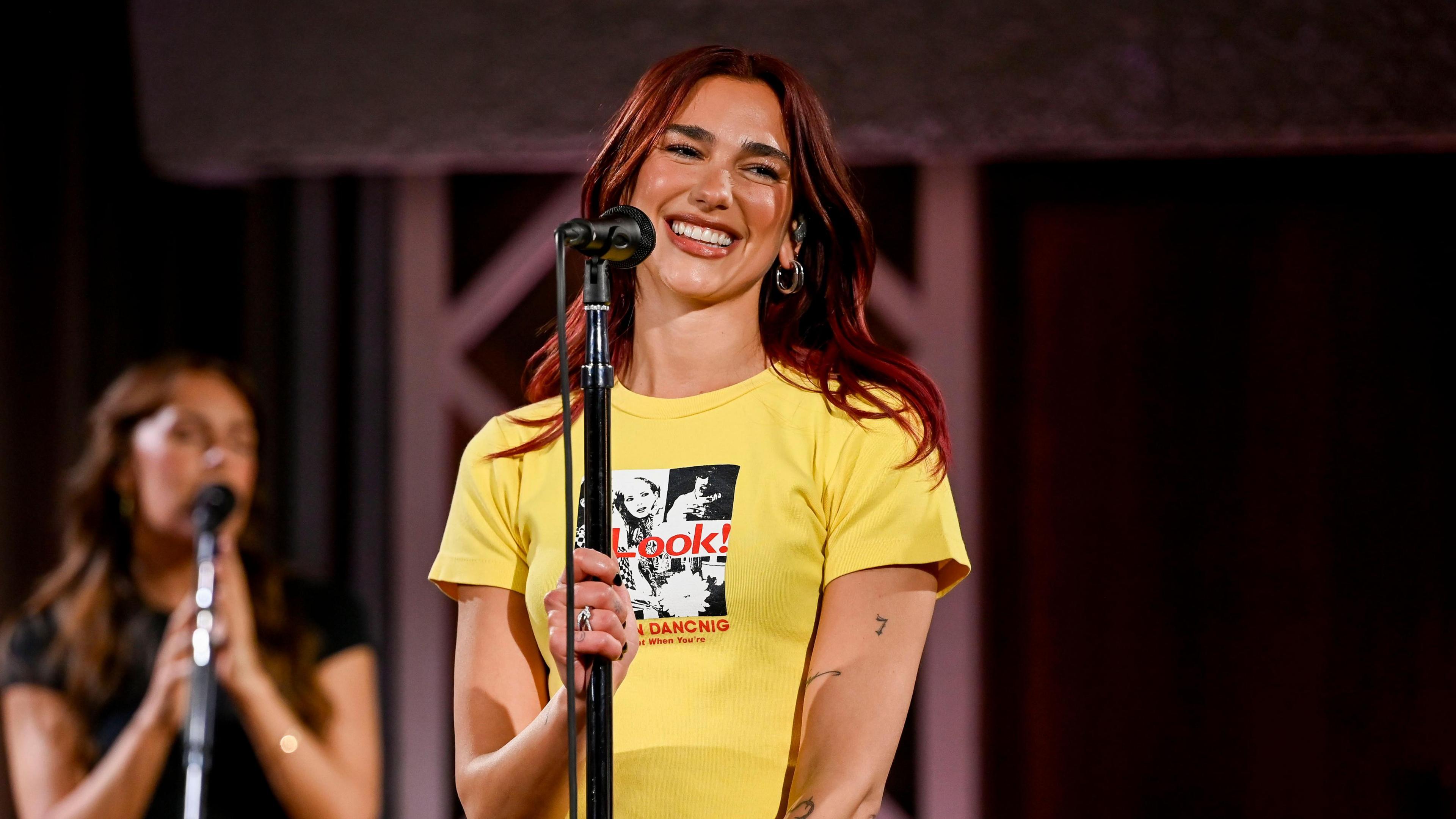 Dua Lipa stands in front of a microphone wearing a yellow t-shirt.