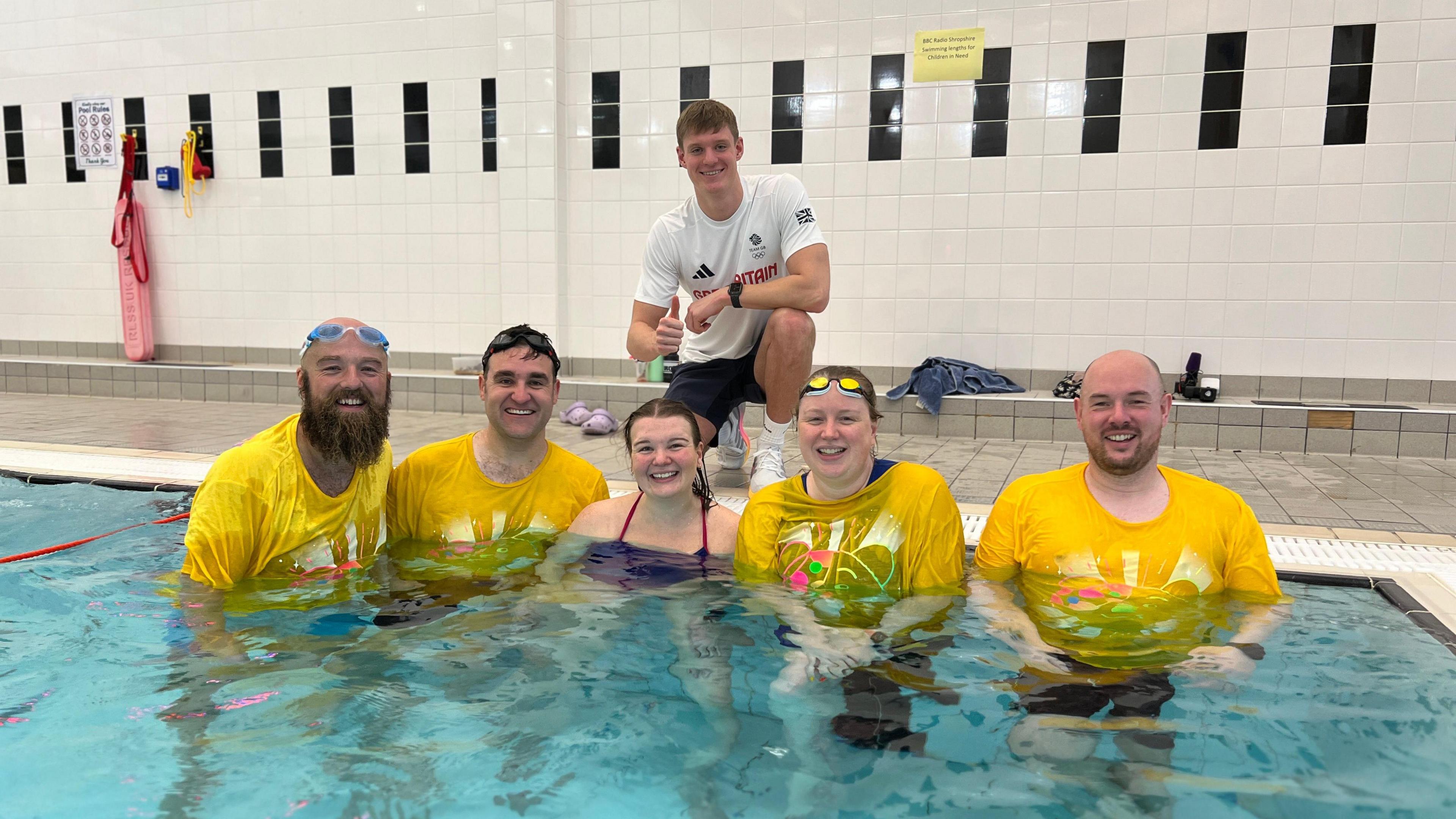 Oliver Morgan is crouching on the side of a swimming pool wearing a white T-shirt. Below him in the pool are five people. Two men on the left are wearing yellow Children in Need T-shirts, a woman is wearing a swimming costume. To her right is a woman and a man, both also wearing yellow Children in Need T-shirts