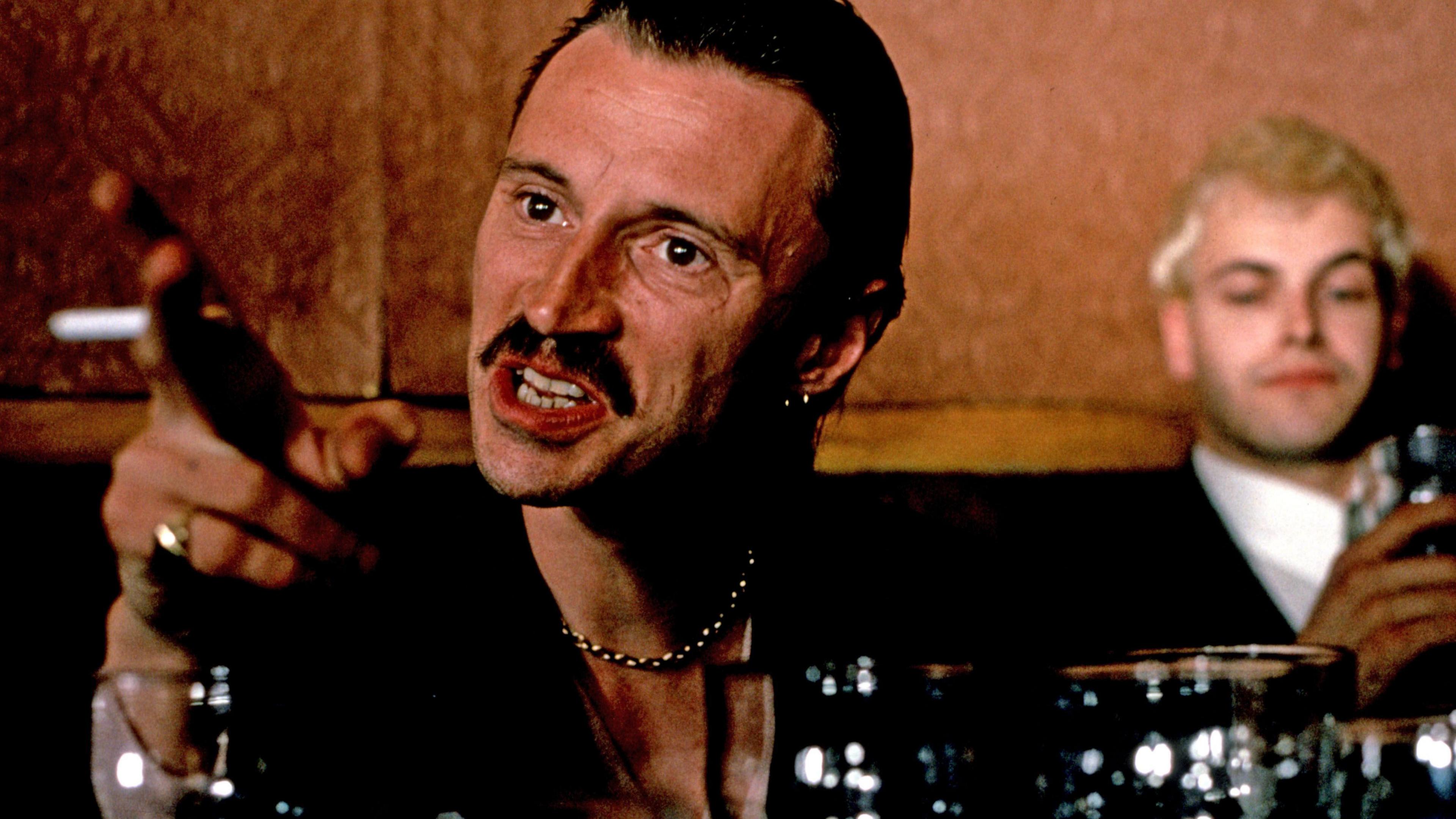 Photo of Begbie in the Trainspotting film. He's in a pub with empty glasses in the foreground. He is sitting at a table pointing menacingly with a cigarette between his fingers. He has a moustache, slicked back black hair and a silver necklace.