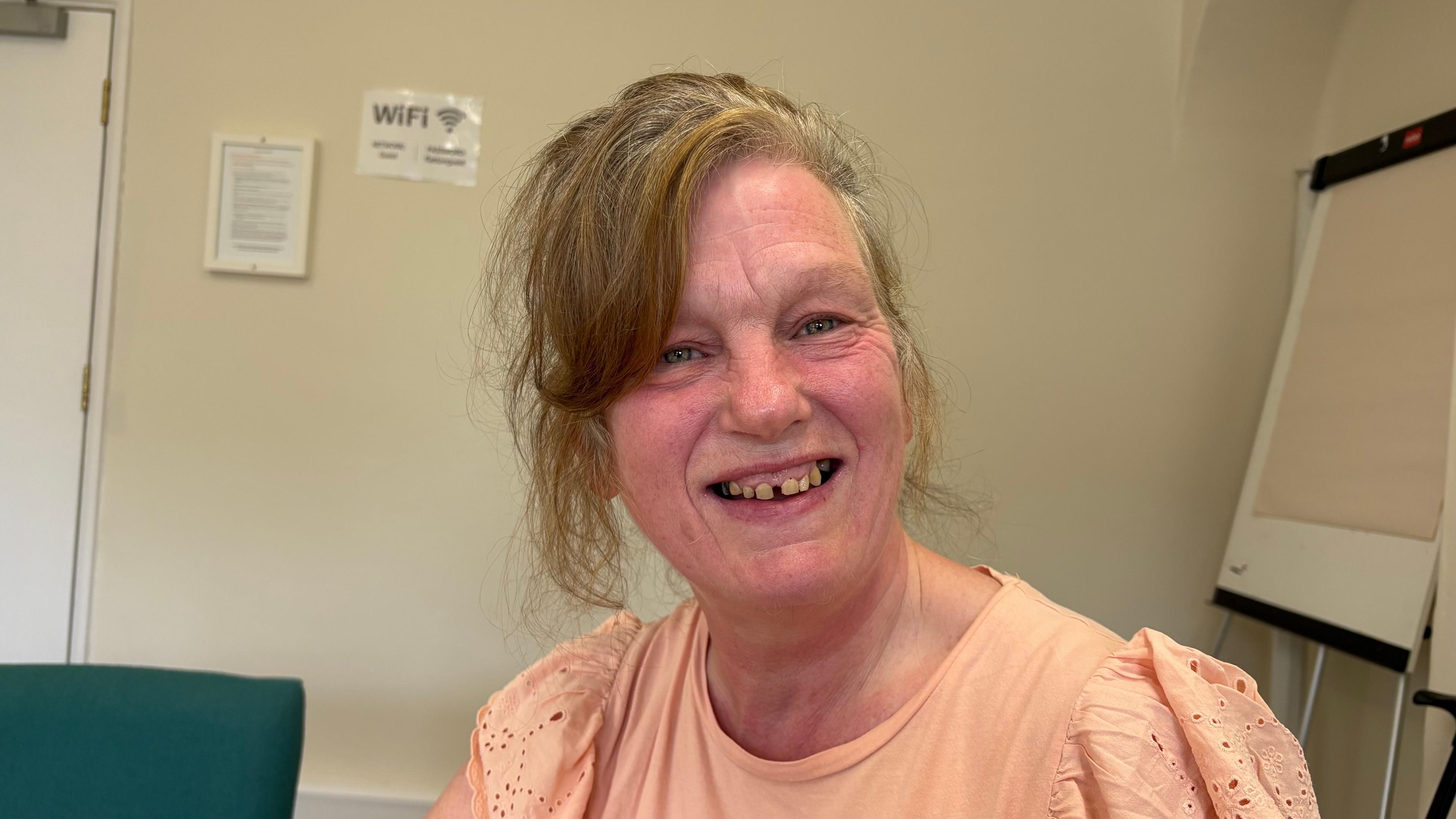 Debbie Flaherty, with long brown hair and glasses, wearing an orange top. She is smiling at the camera with a light-coloured wall behind her. There is a flipchart leaning against the wall to the right.