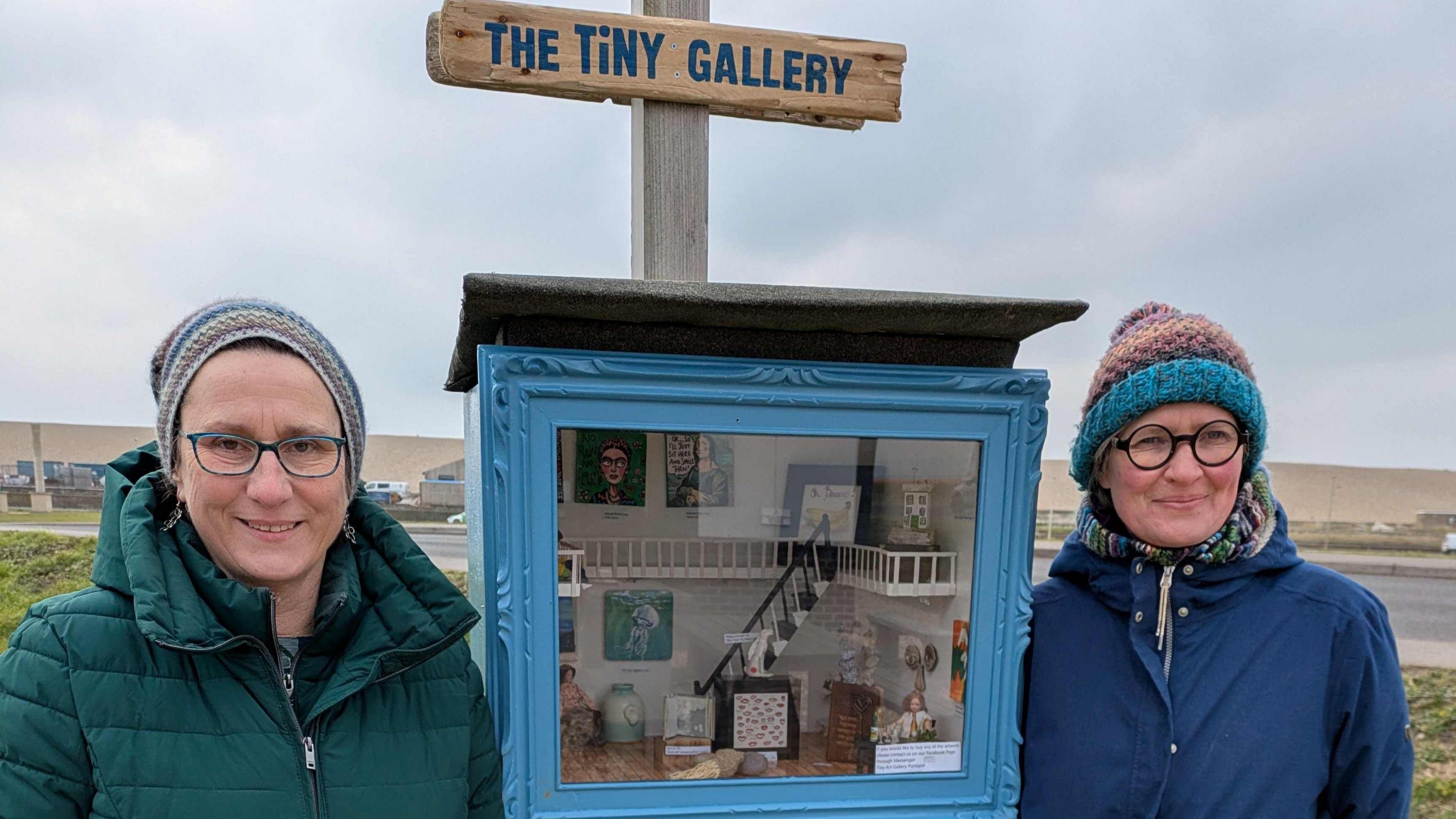 (Left to right) Antje Rook smiling at the camera and wearing a green puffa jacket and beanie hat and Yollande Posthumus is also smiling at the camera wearing a blue raincoat and a knitted hat. In between them is a beehive box where the tiny gallery can be viewed.