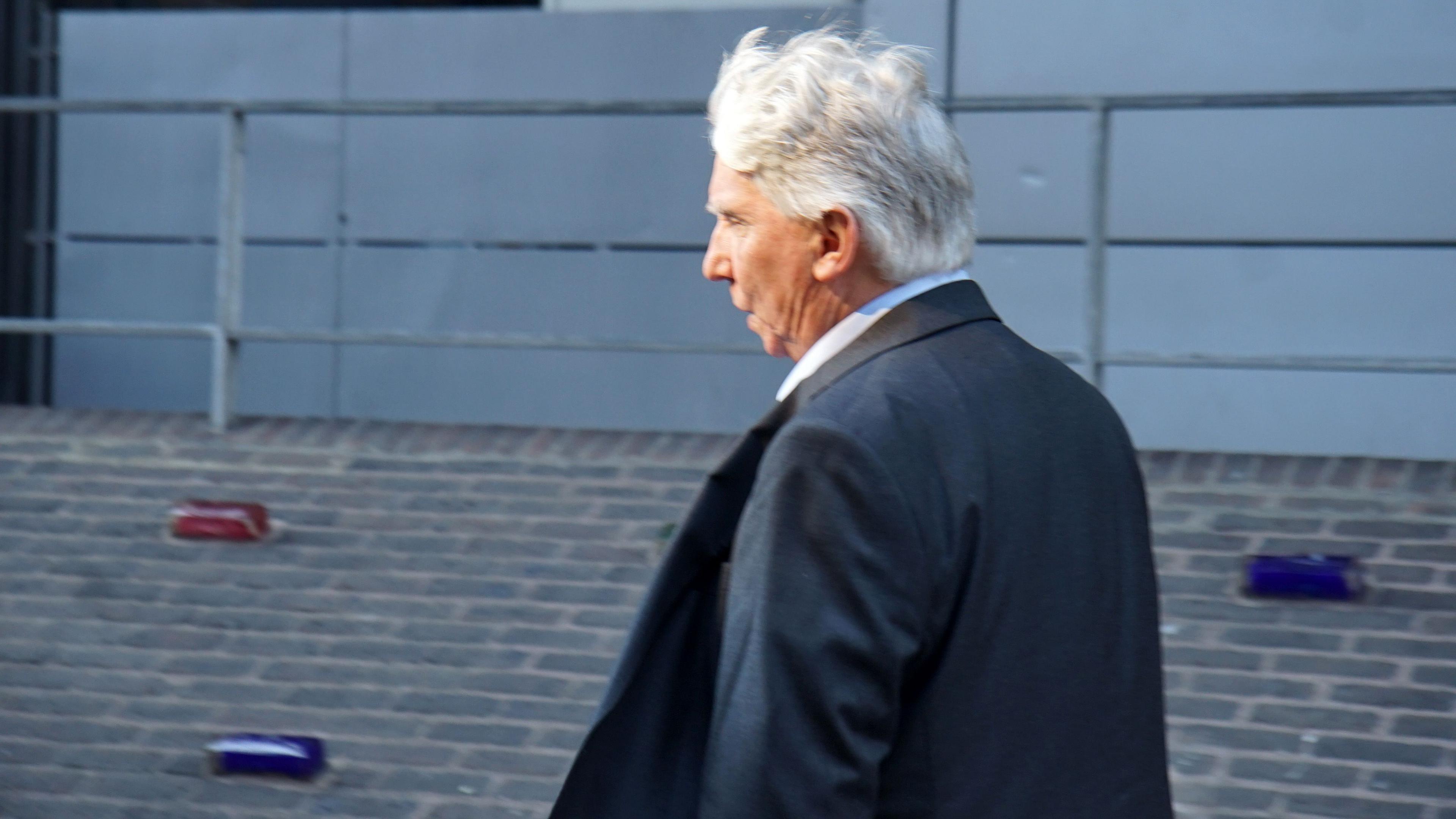 David Geary, a man with gray hair, dressed in a dark suit and white shirt, is walking along a brick pathway. Behind him, a metal railing runs parallel to the path, and the background features a gray wall with horizontal lines, giving the scene a modern, urban appearance.