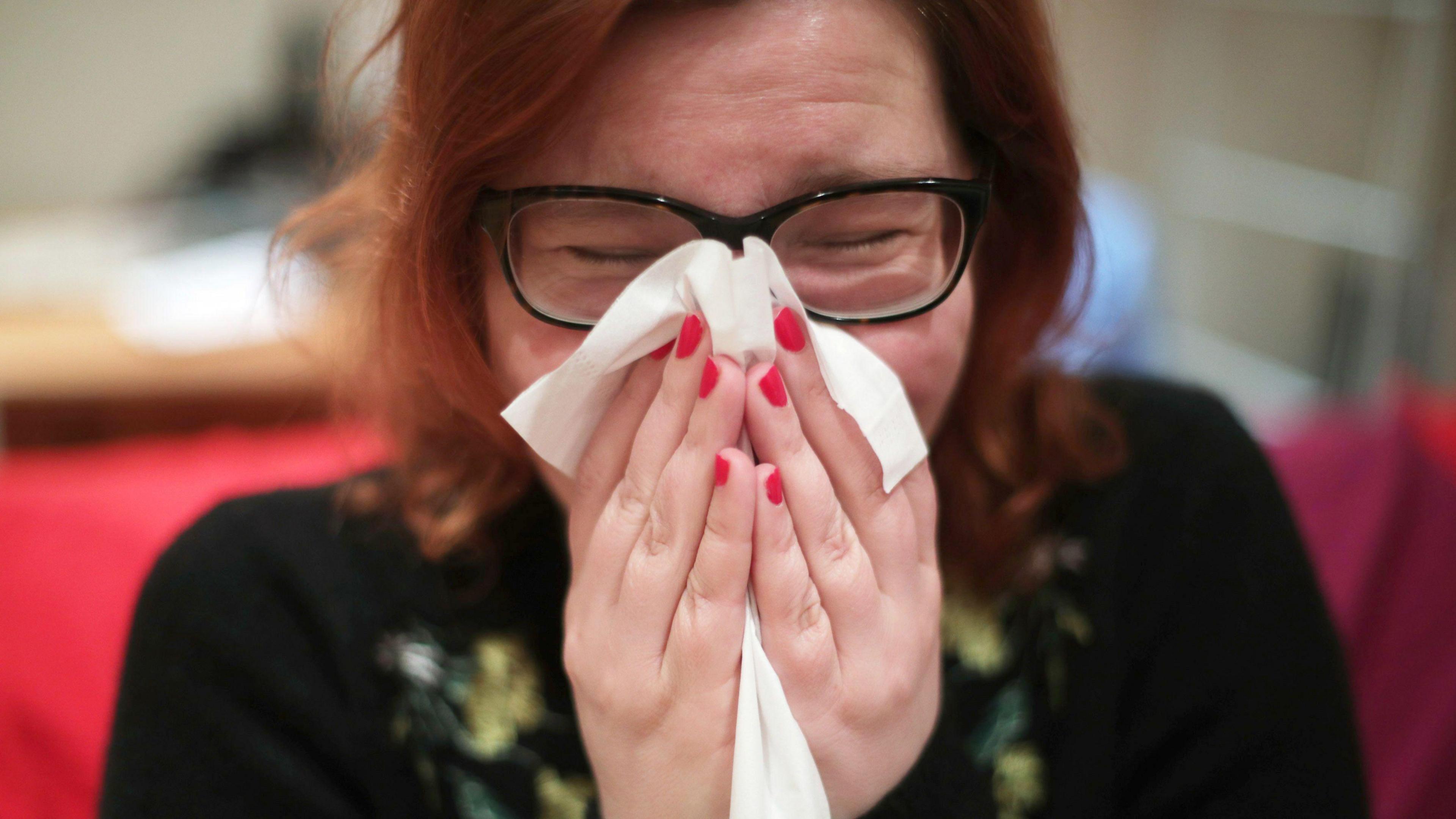 A close-up of woman with red hair and red painted nails blowing her nose with a tissue. Her eyes are shut behind dark-framed glasses. A red sofa and wooden desk are blurred behind her.