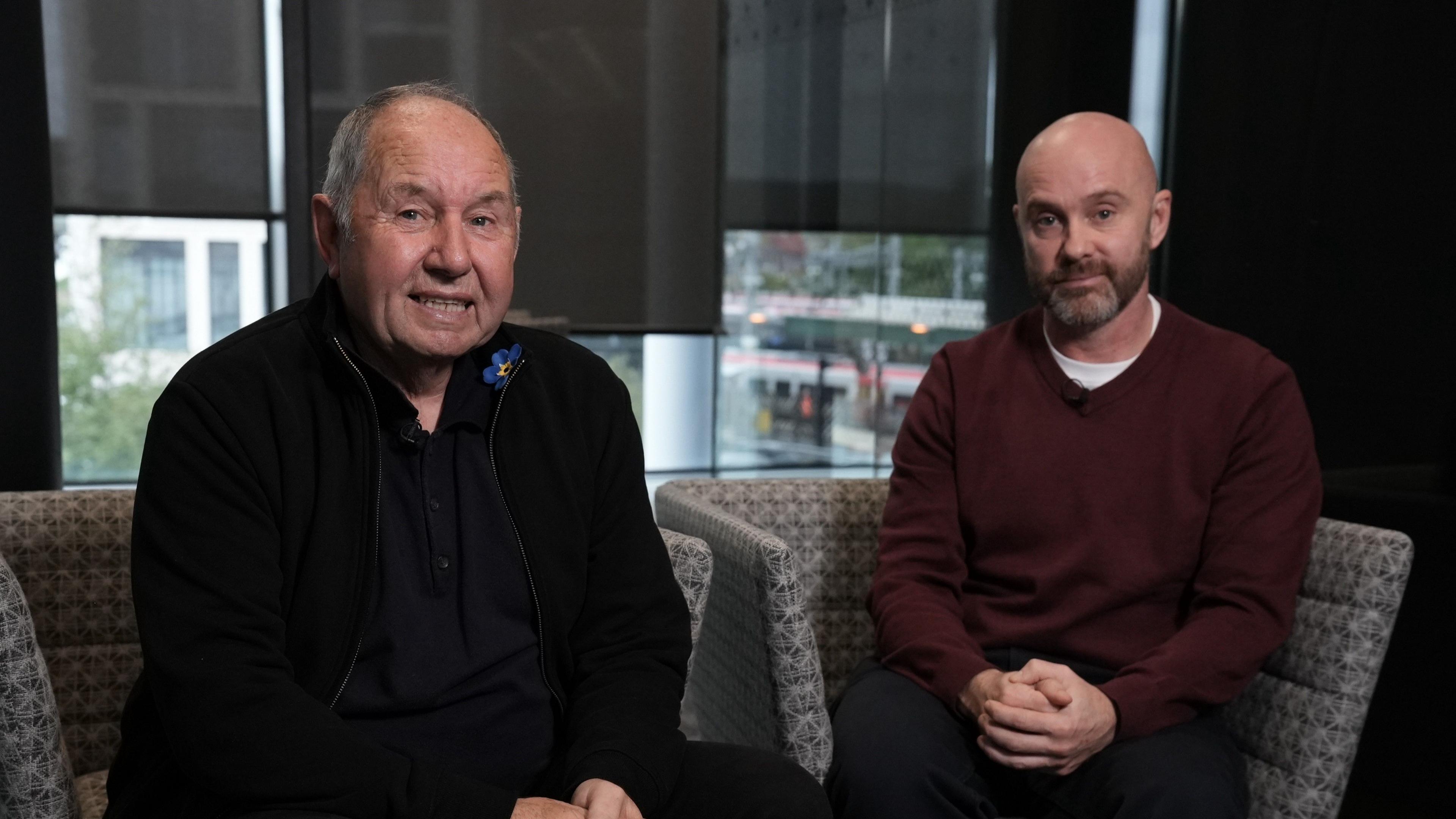Chris Griffiths a Lee Griffths yn gwenu tuag at y camera. Mae Chris yn gwisgo siaced ddu a chrys du gyda bathodyn cymdeithas Alzheimer's ar ei goler. Mae Lee yn gwisgo siwmper coch tywyll.