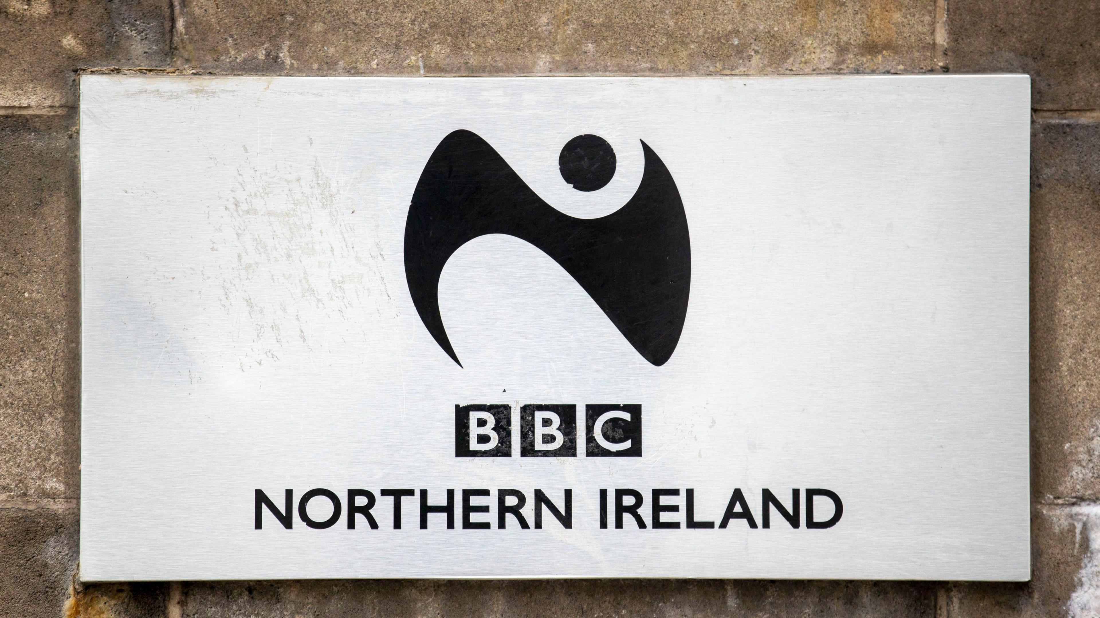 A silver plaque states BBC Northern Ireland with a black logo of a swirl and a circle. It's attached to a brick wall.
