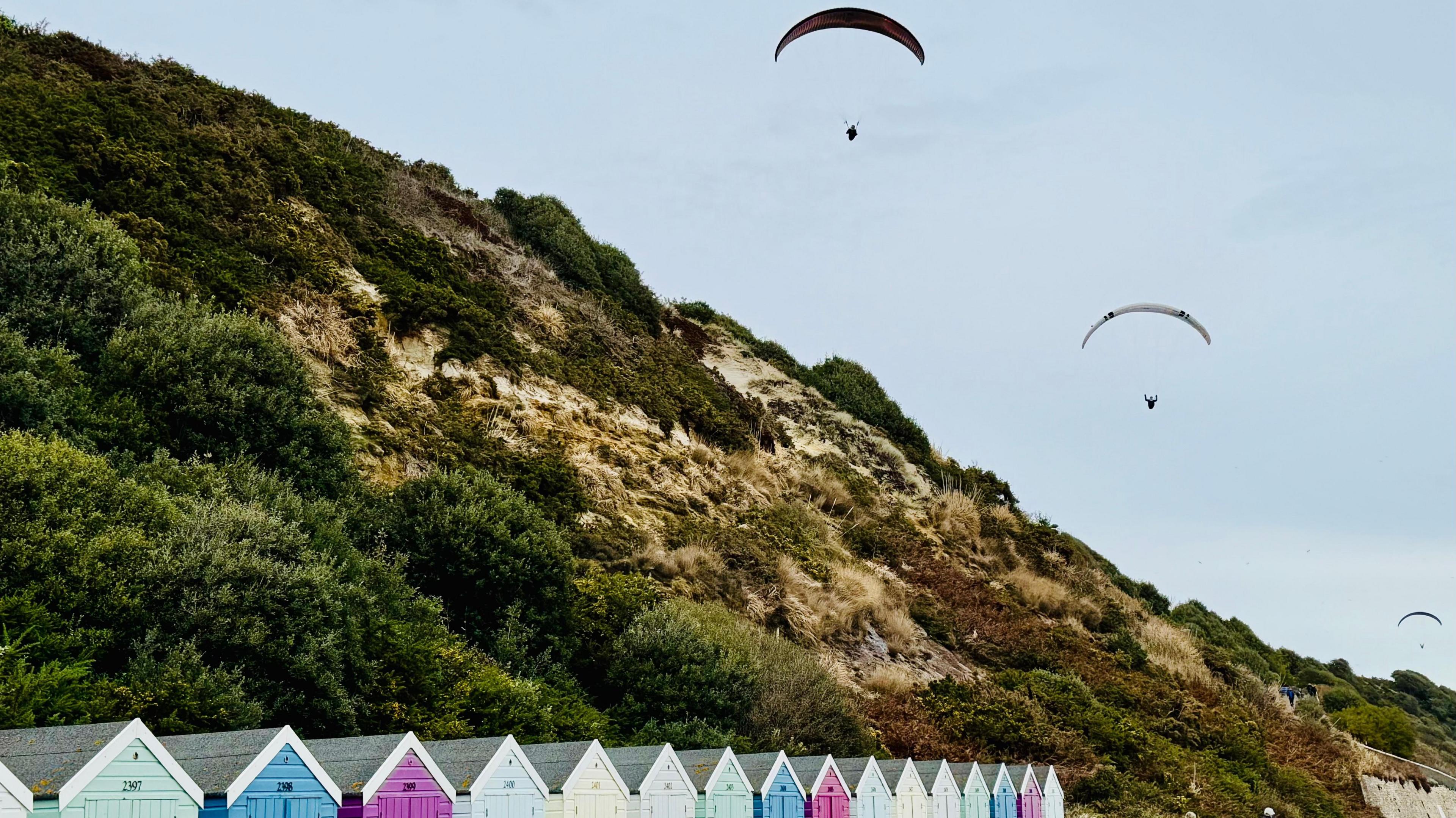 Three paragliders line up in the sky with arched canopies above beach huts and the gently sloping, vegetated cliff at Boscombe.