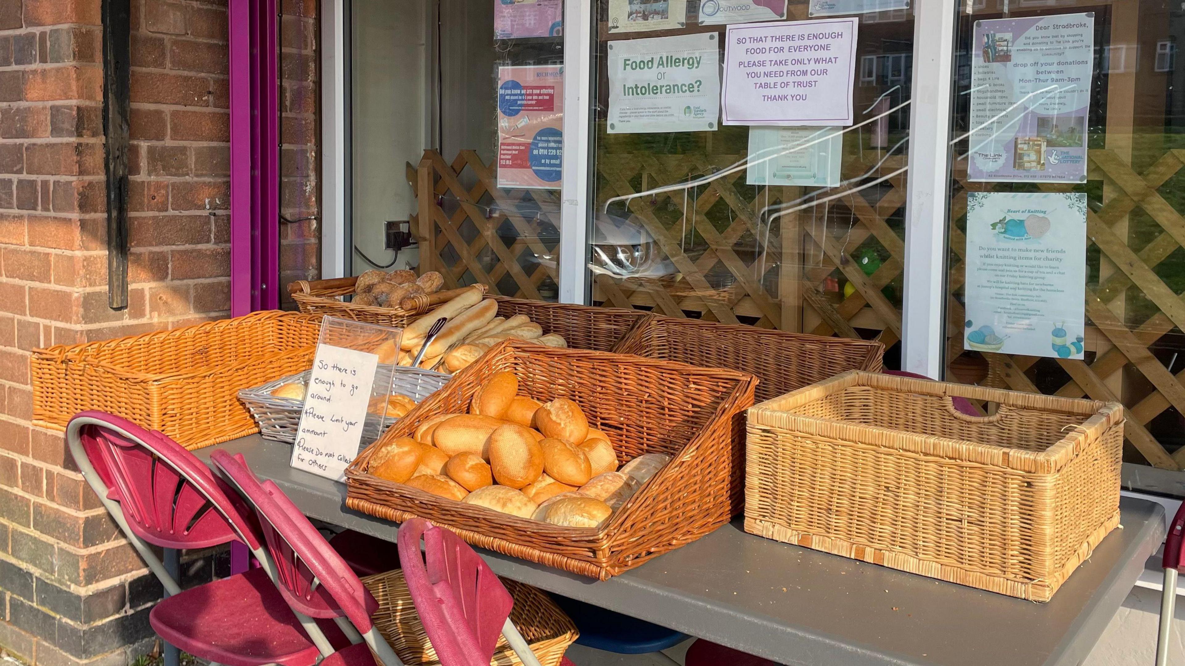 Baskets of bread outside the front of the shop. There are bread rolls and baguettes. A sign tells people to limit the amount of food they take, so there is enough for others.