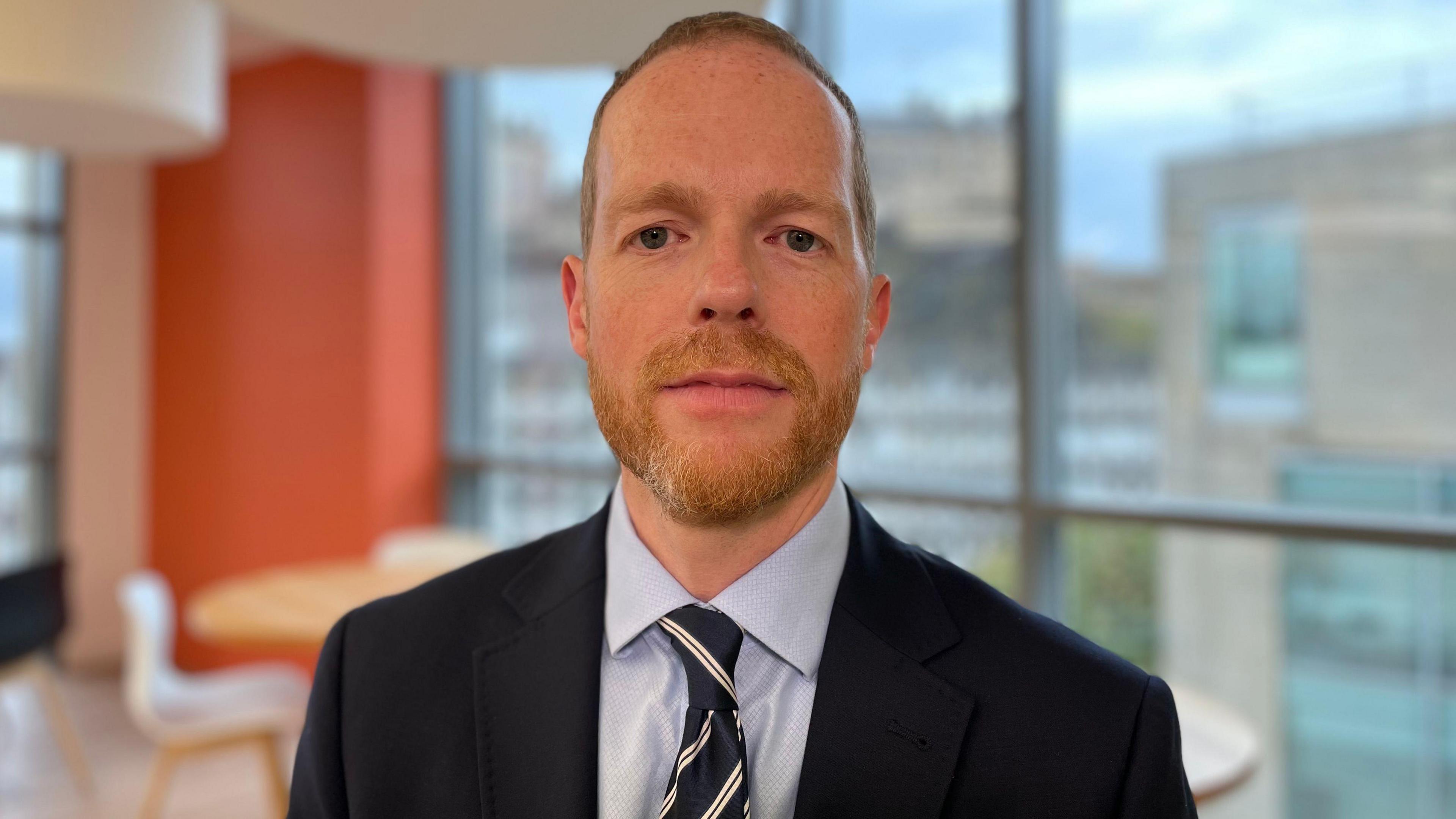 Auditor General for Scotland Stephen Boyle photographed from the chest up in an Edinburgh office. He has short red hair and a trimmed beard and moustache. Mr Boyle, who is staring straight at the camera, is wearing a navy suit, light blue shirt and a navy, white and red striped tie.