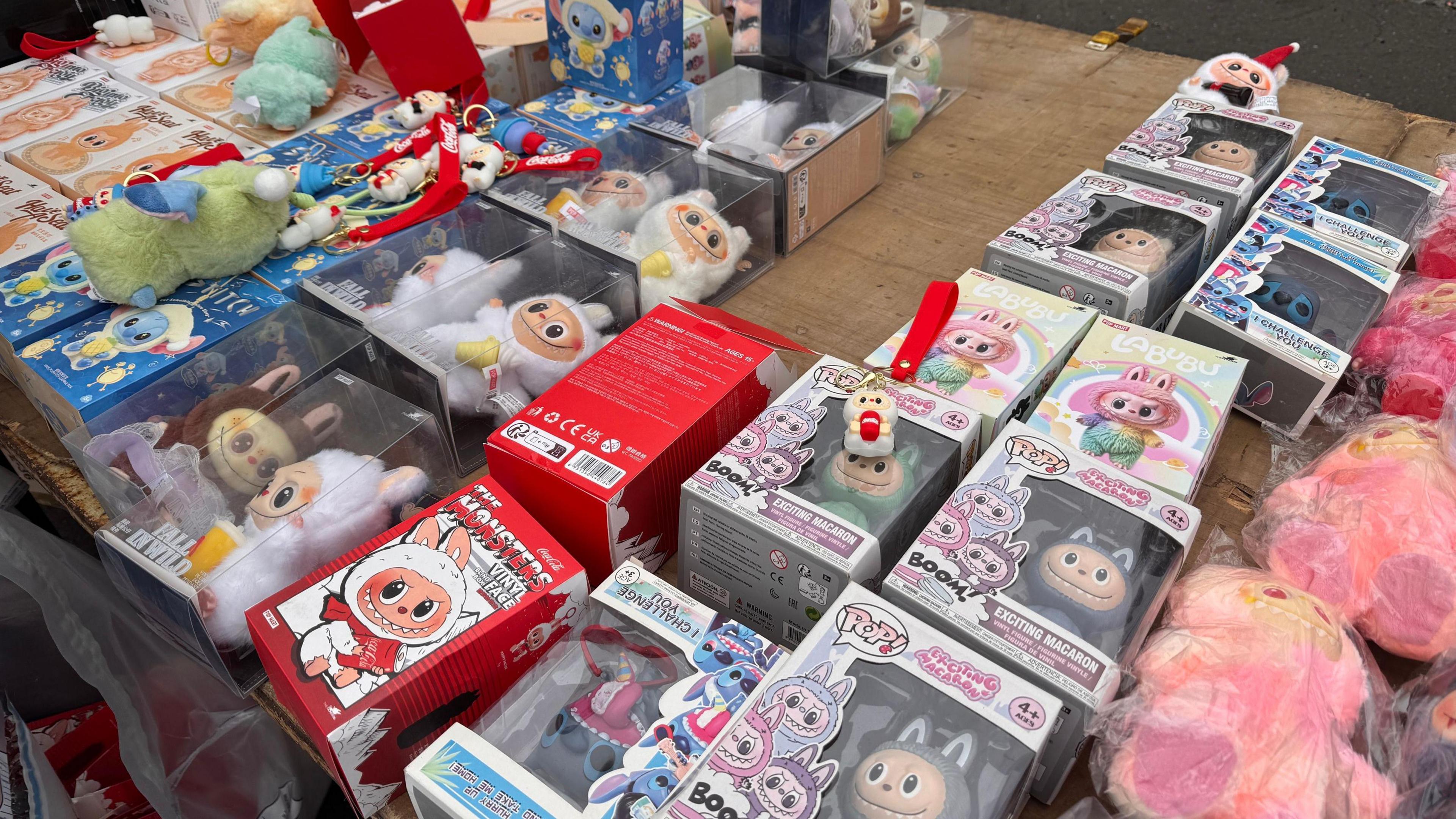 A number of fake Labubu dolls of different colours displayed on a market stall table in various colours and boxes.