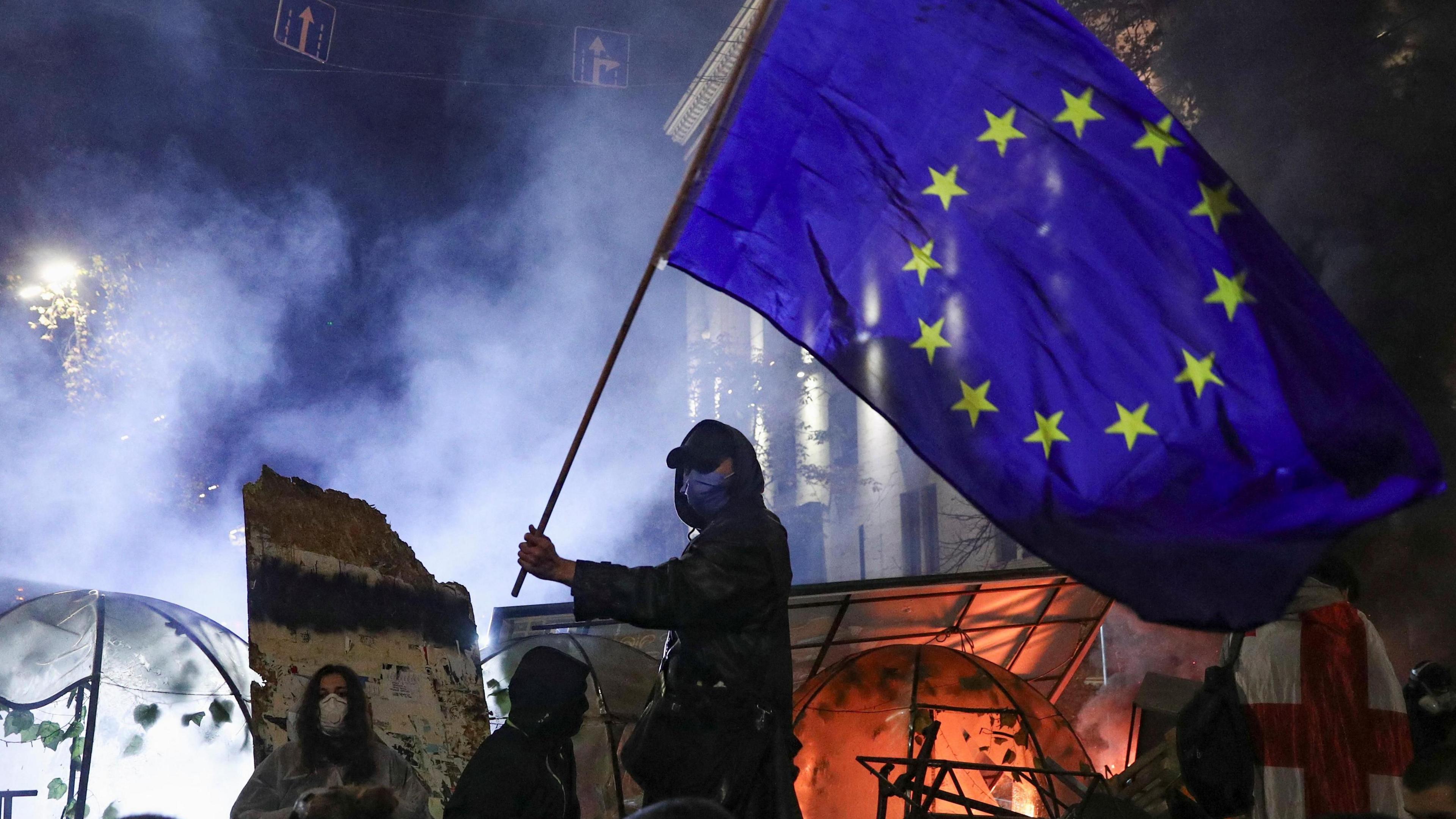 A masked protester waves an EU flag above a makeshift barricade at the protests. There's smoke in the background.