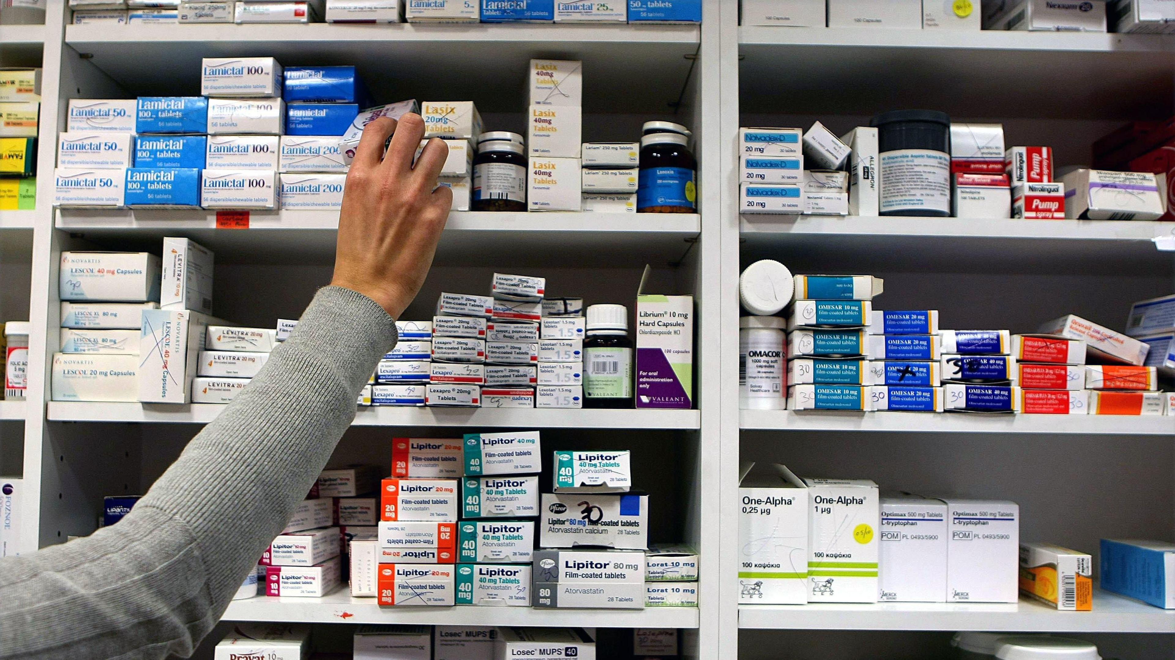 A close-up image of a hand grabbing a small box of prescription drugs from a shelf. The shelves around are full of stacked small boxes with multi-coloured labels.