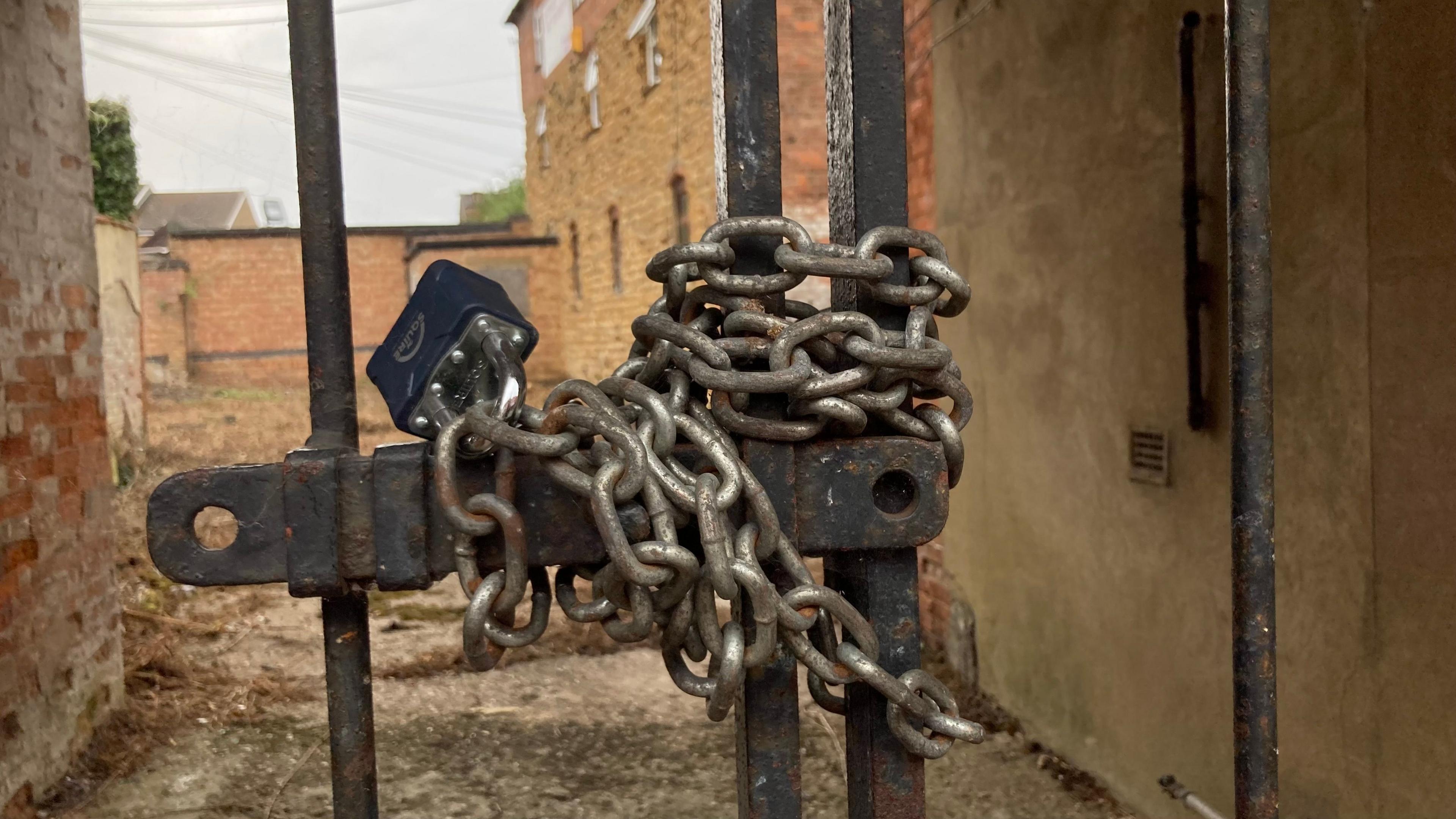 Padlocked chain wrapped around an iron gate. A derelict building and yard are visible behind.