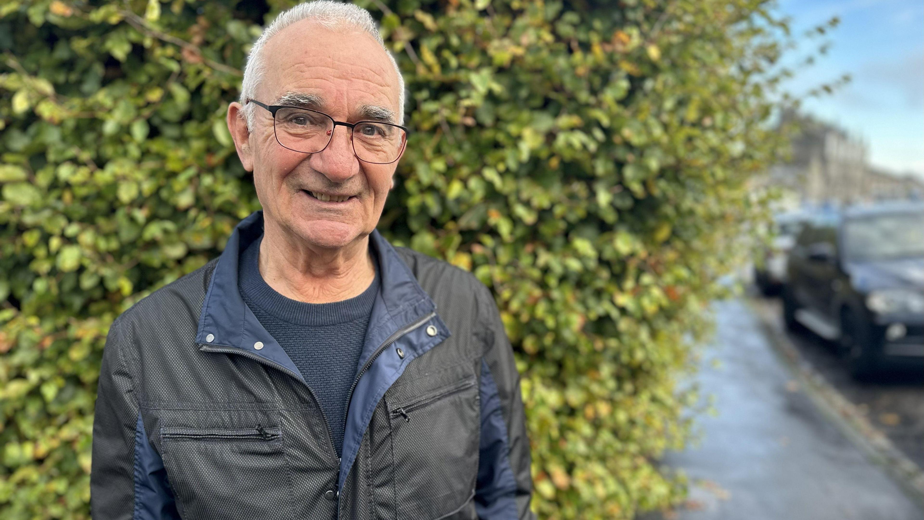 Jim Gordon standing outdoors on a wet pavement beside a leafy green hedge. He is wearing a dark jacket with a zip-up front and layered panels, along with a dark sweater underneath.