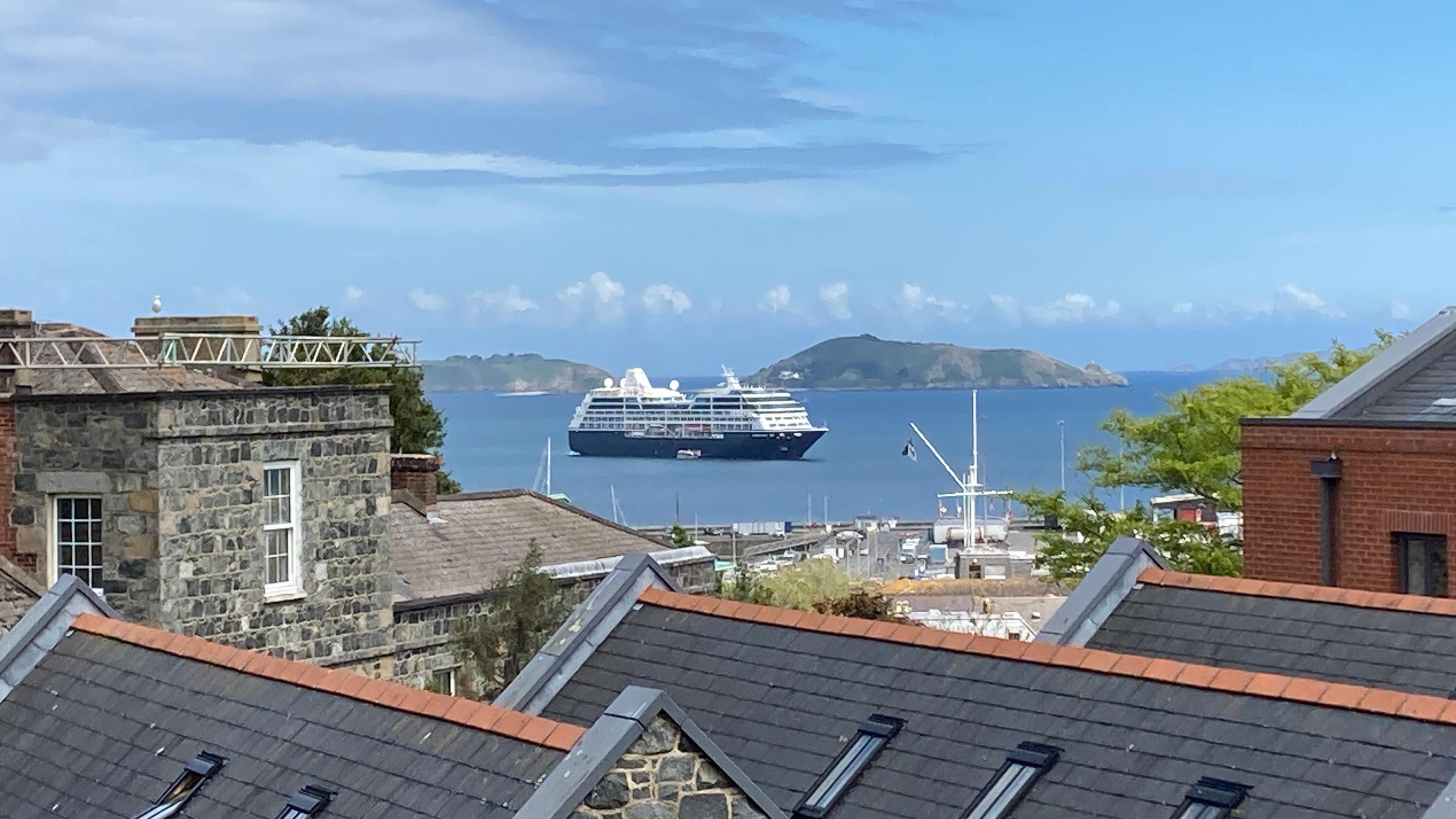 A view of a white and blue cruise ship in water between Guernsey and Herm. Houses and a church are in the foreground 