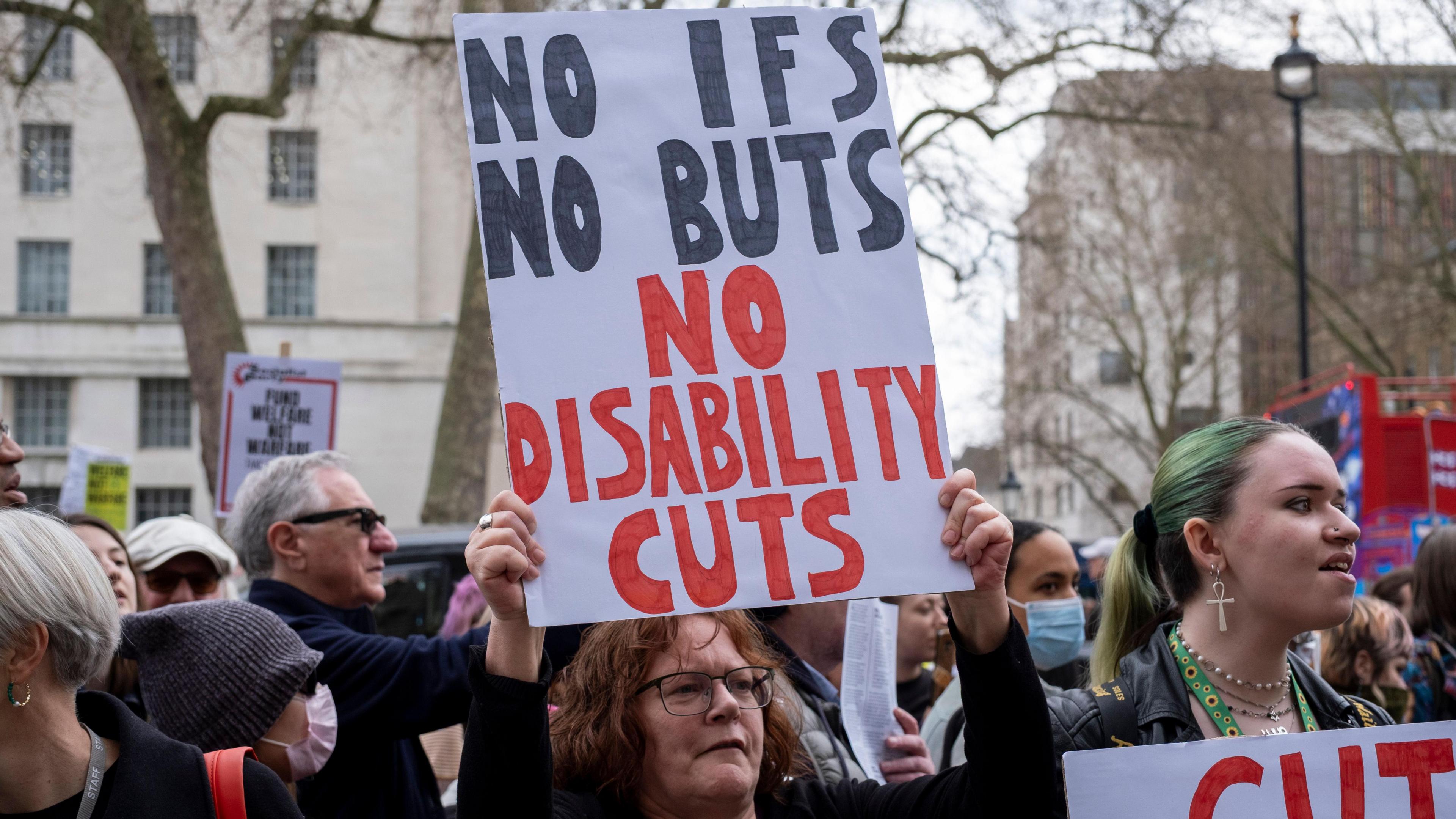 A protester holding a sign reading "No Ifs, No Buts, No Disability cuts"