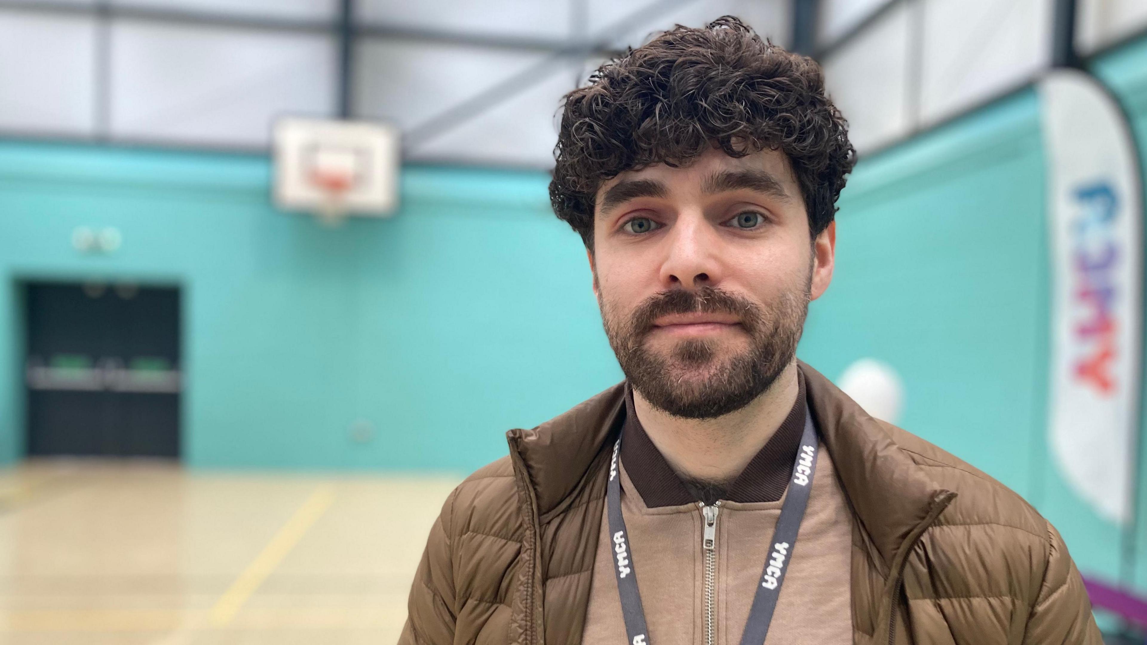 A young man standing in a sports hall looking towards the camera. He has a beard and dark-brown curly hair and is wearing a brown padded jacket. He has a black YMCA ribbon around his neck