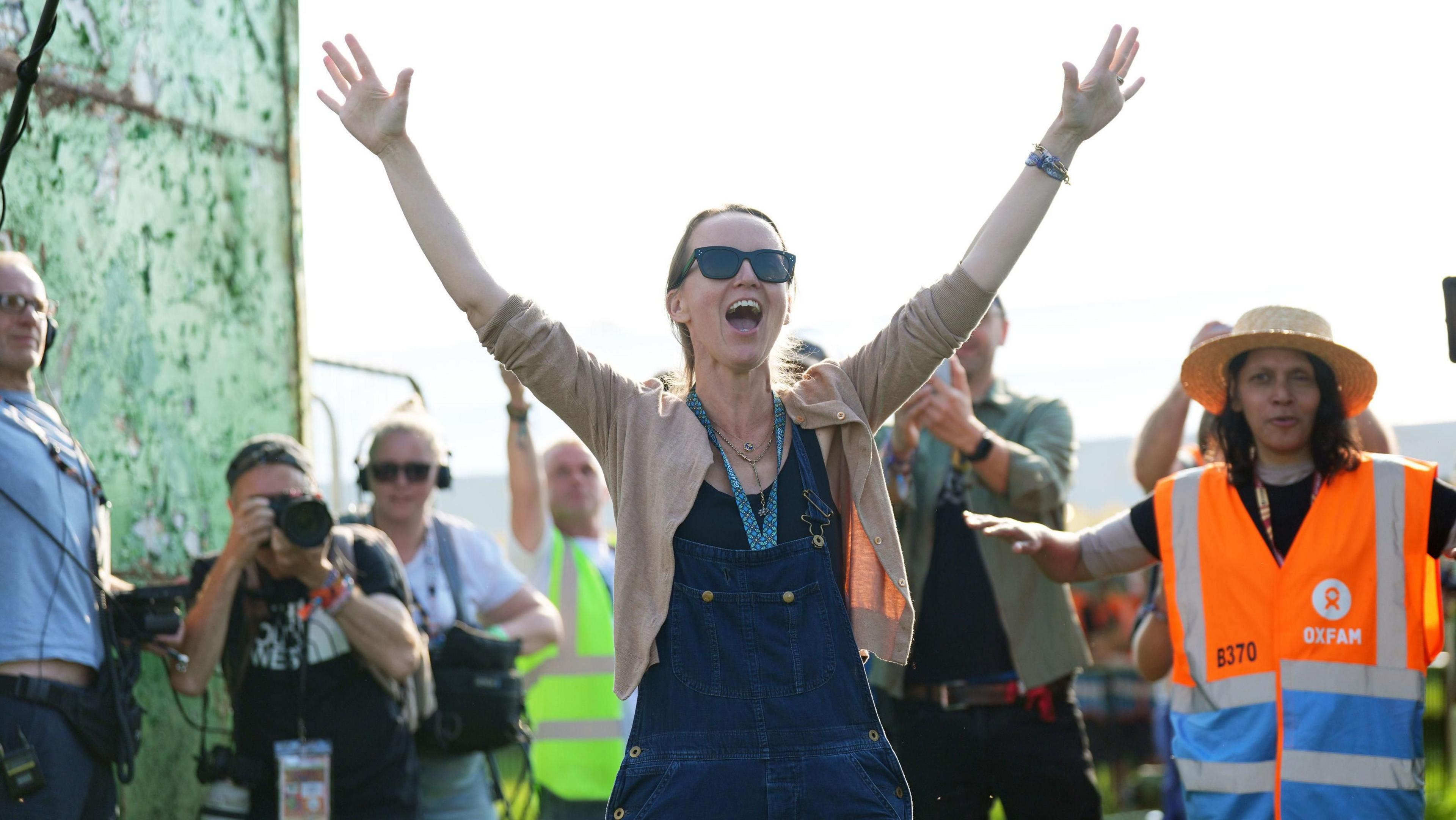 Emily Eavis standing in front of green gates, with photographers and security staff in high viz jackets behind her. Emily has her arms in the air.