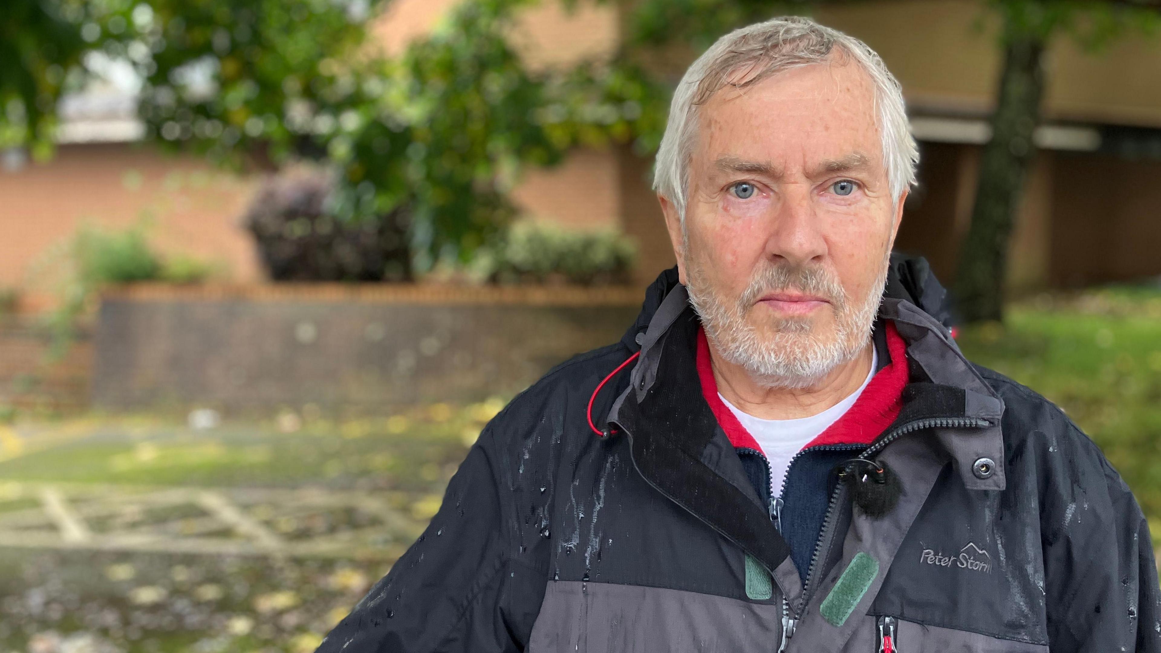 Hugh Janes in front of the registry office building. He has short grey hair and a short grey beard. He is wearing a grey raincoat and a blue and white fleece. 