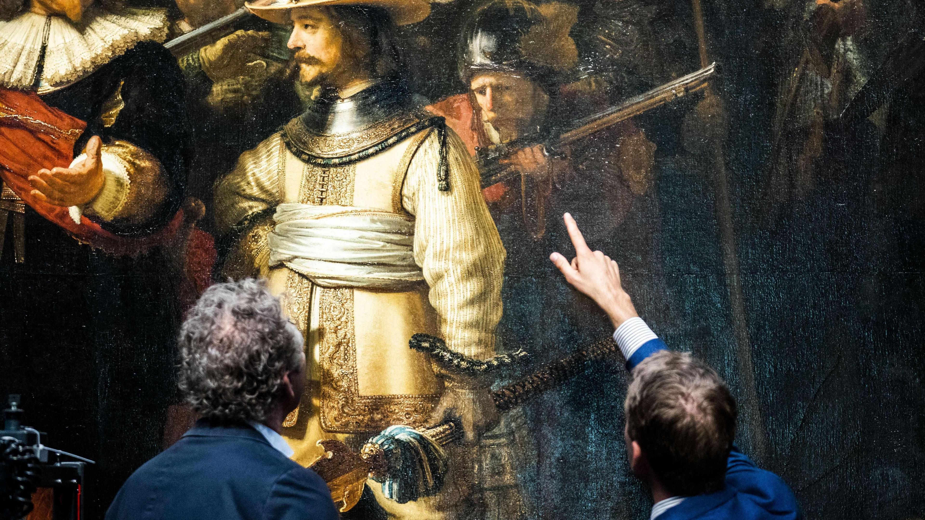 Two visitors in front of Rembrandt's The Night Watch. The visitor on the right is pointing at lieutenant Willem van Ruytenburch in the painting, who is dressed in a gold outfit with a white sash. 