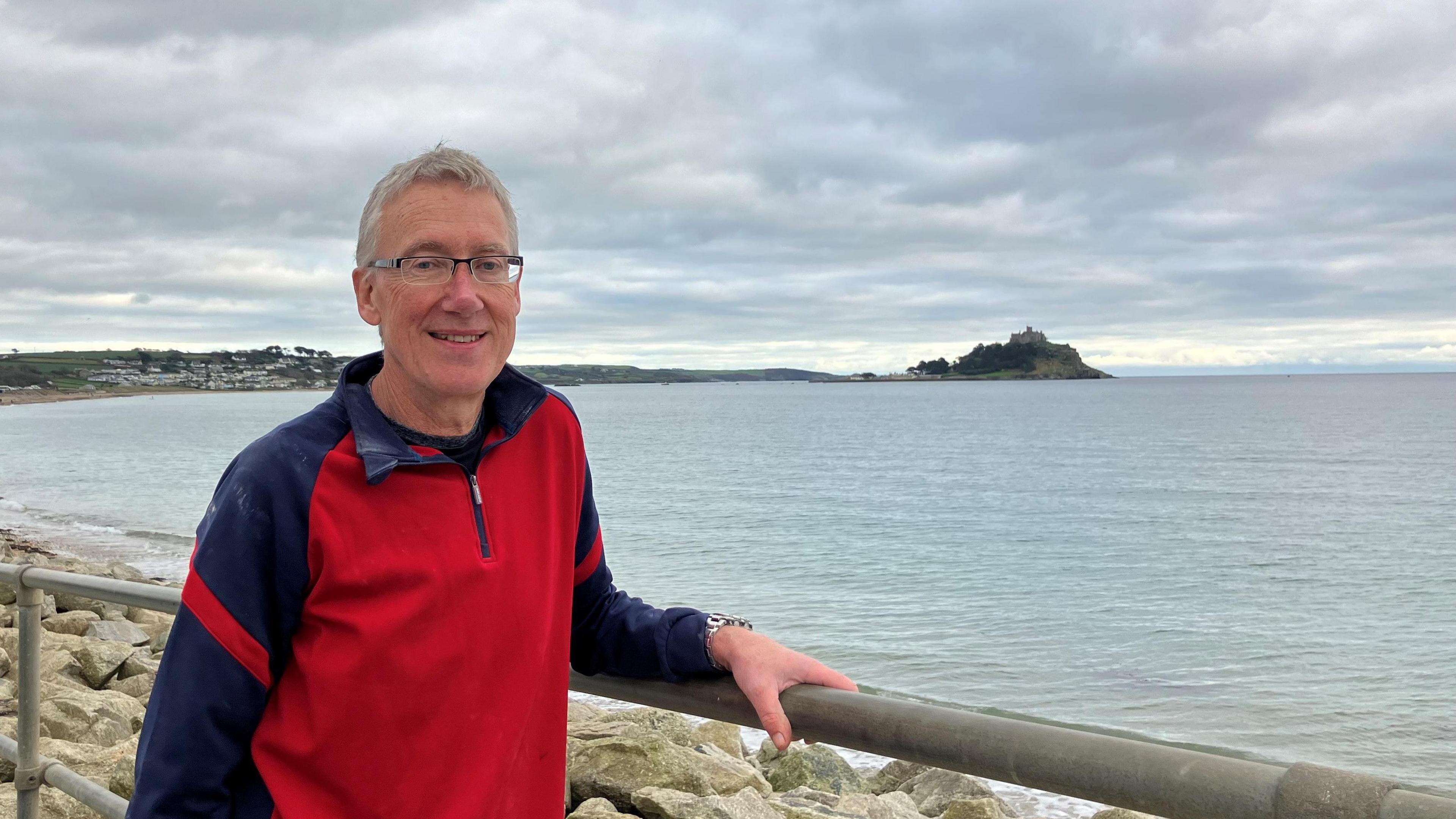 Simon Ayres, in a red and blue jumper, is smiling and standing in front of the sea with St Michael's Mount in the background.