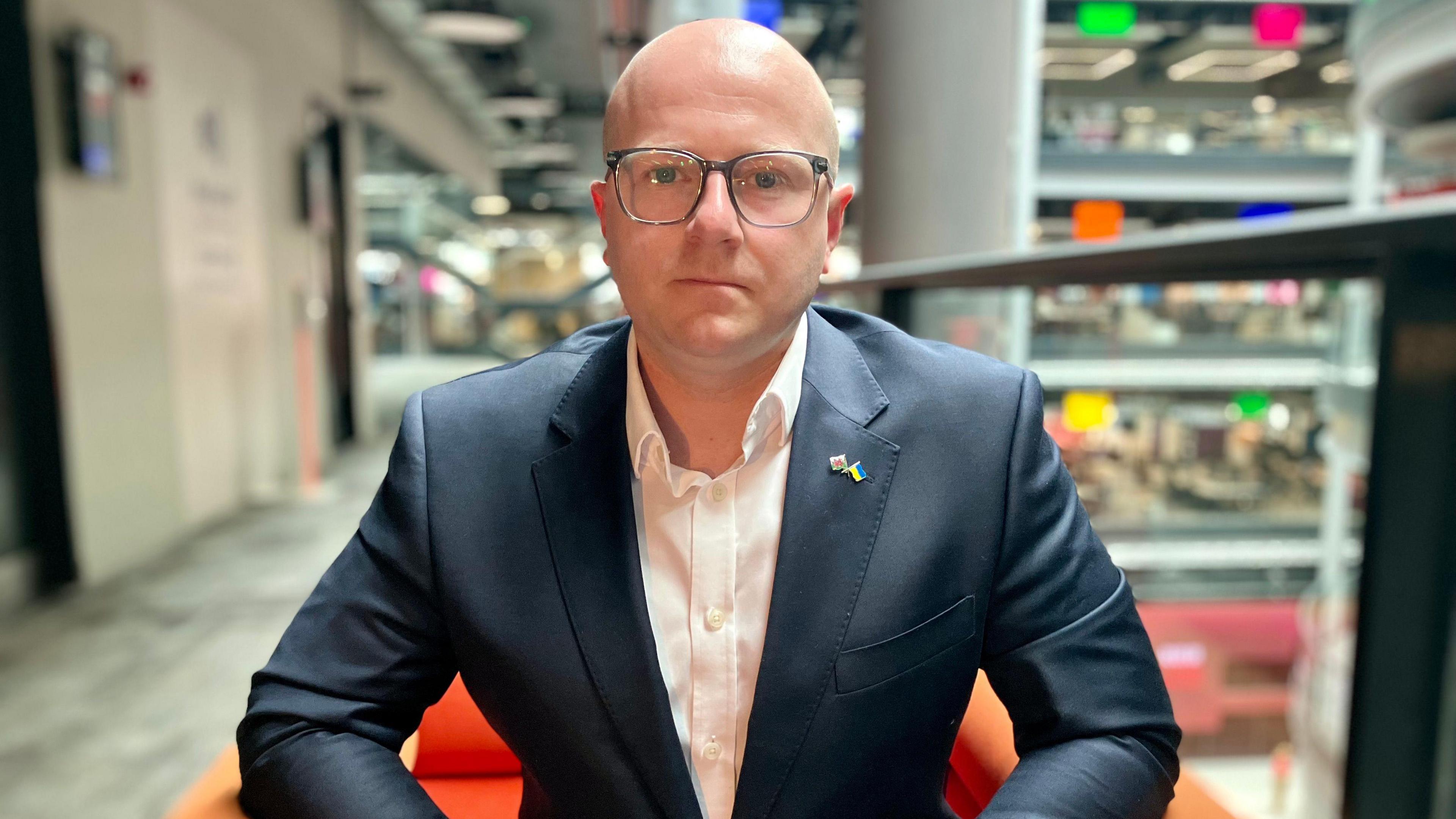 Carwyn Donovan has a bald head and is wearing glasses. He is wearing a dark suit with a Ukraine and Wales flag pin badge. He is also wearing a white open-necked shirt