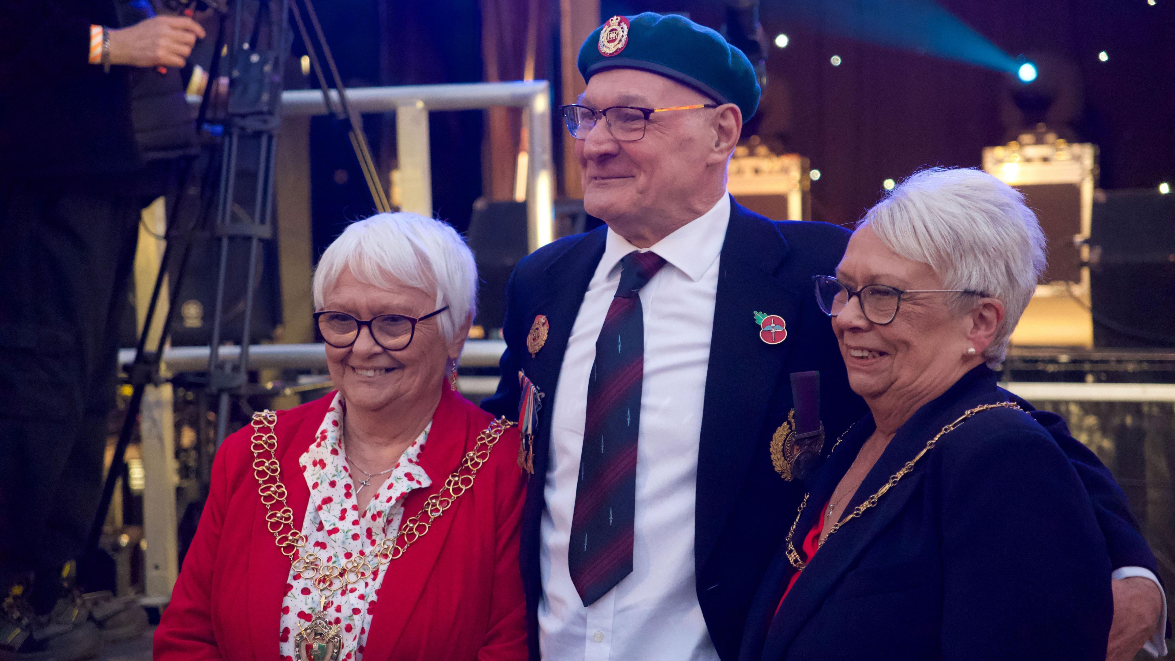 A woman in a red jacket and red spotted white dress, wearing a mayoral chain. Next to her is a former serviceman with beret, blue blazer and navy and purple tie. On the right is a woman with a blue jacket and civic chain