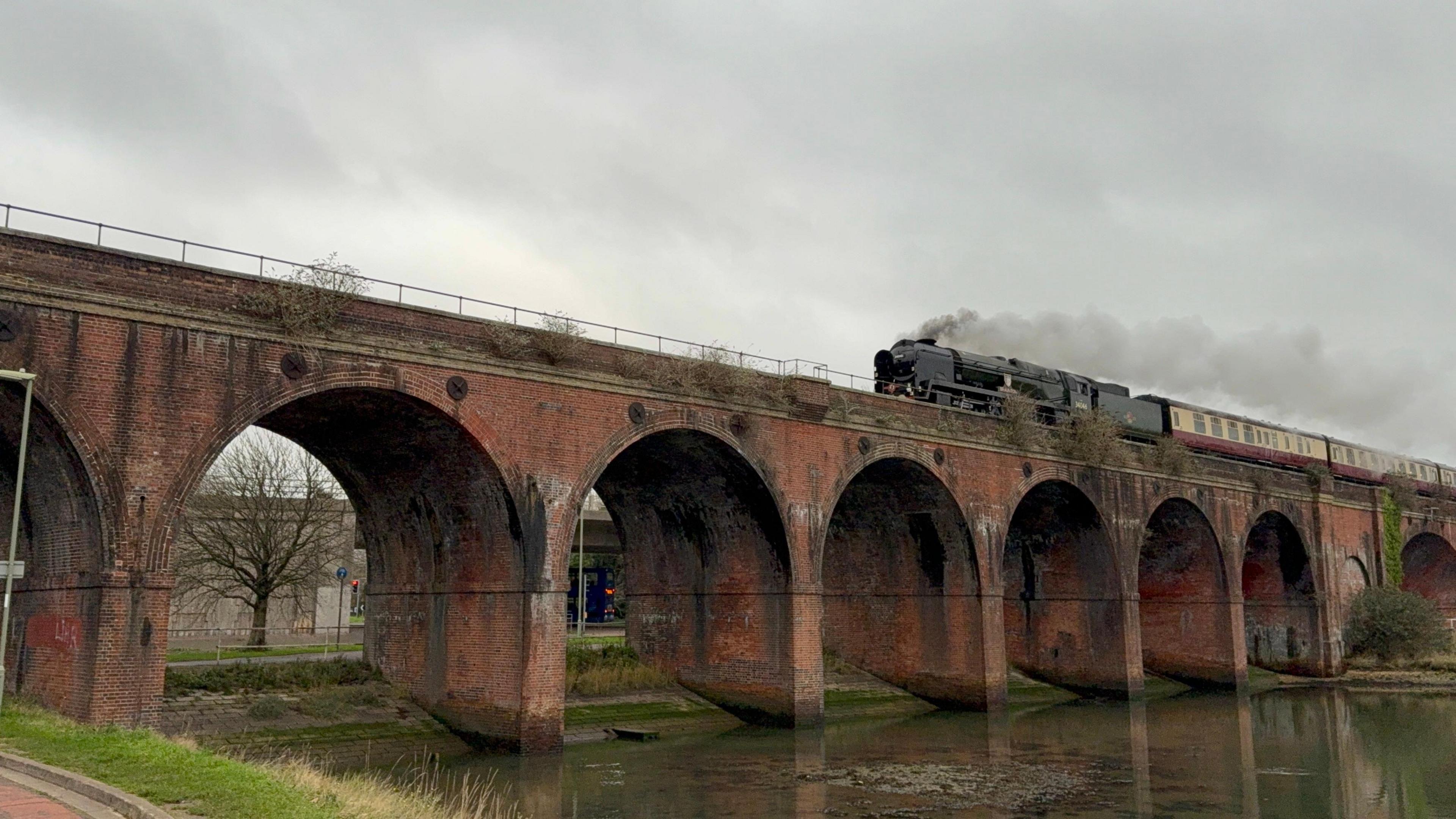 A bridge spans a river and is made up of six arches. A train is just coming over the bridge with steam coming out of its chimney. It is pulling several carriages that are yellow and red.