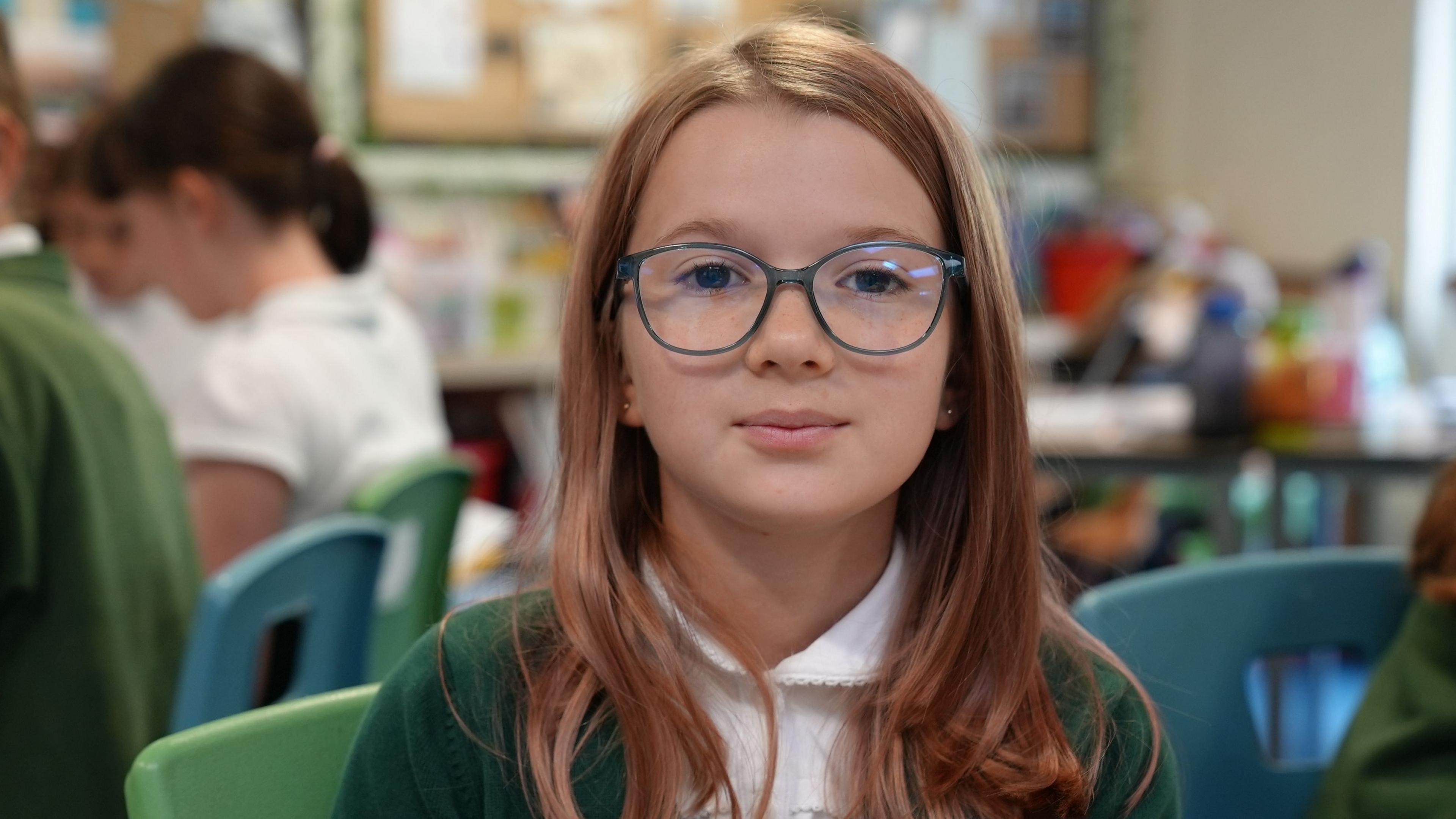 A school girl with long brown hair and blue rimmed glasses looks into the camera 